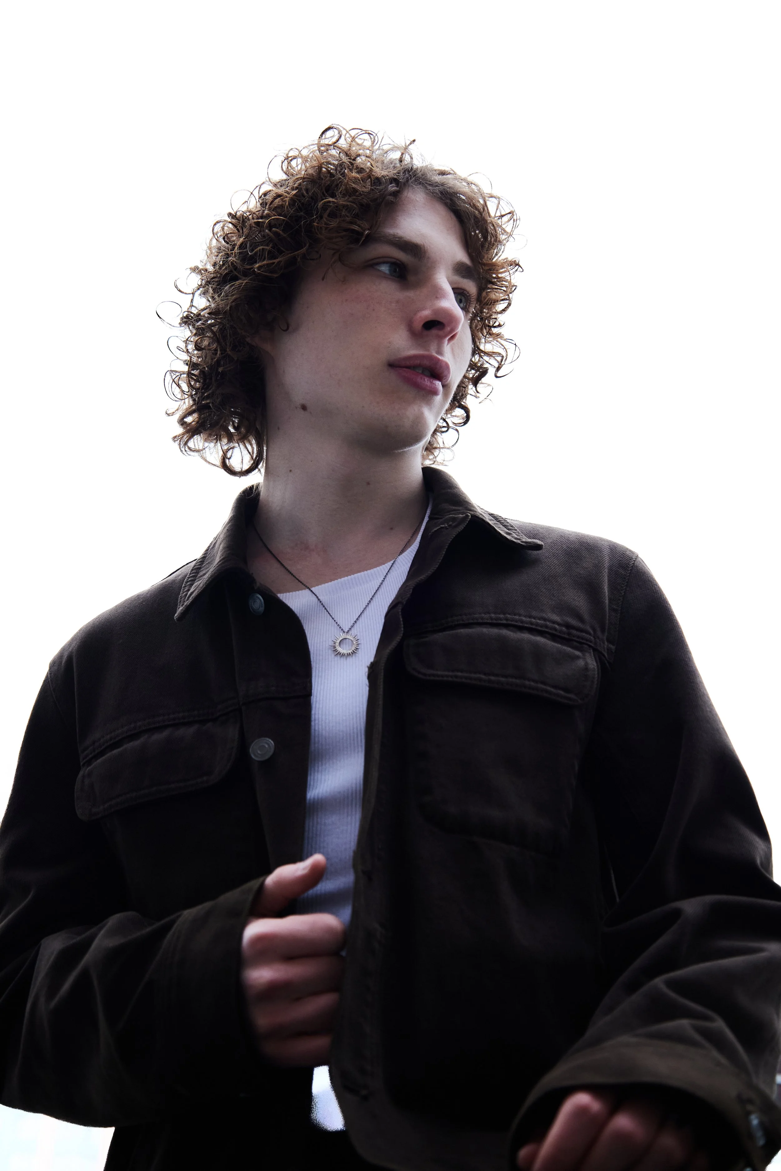 A young person with curly hair looking off to the side, wearing a black jacket over a white shirt, and a necklace with a circular pendant, against a white background.