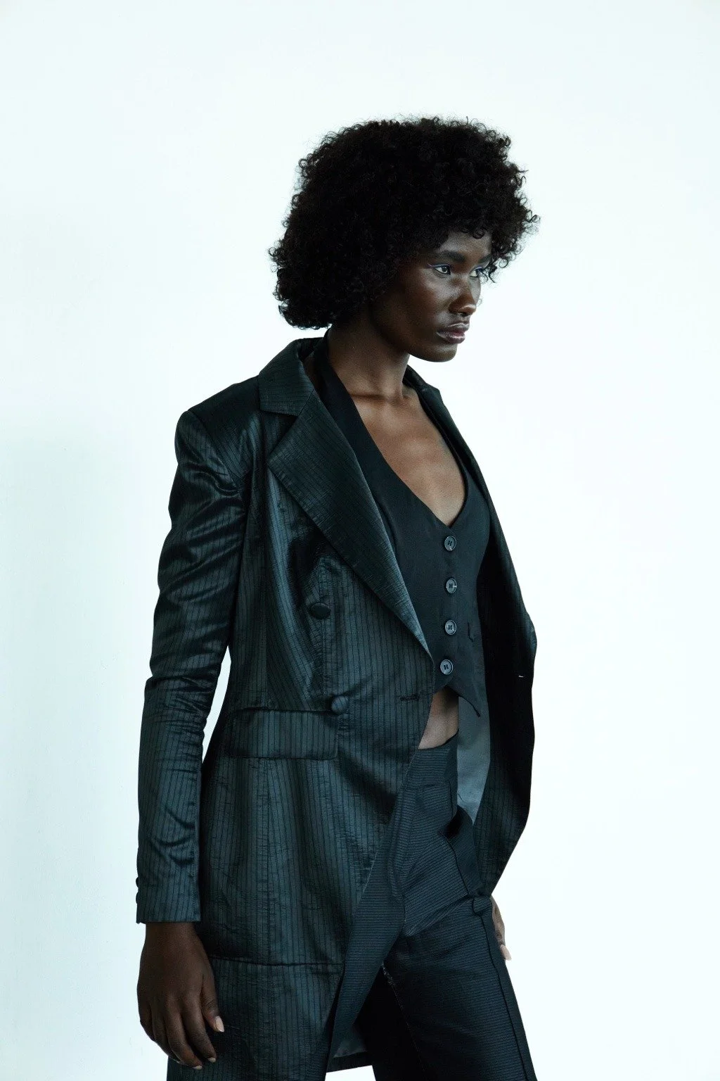 A woman with dark skin and curly black hair wearing a black pinstripe suit and black top, standing against a plain white background.