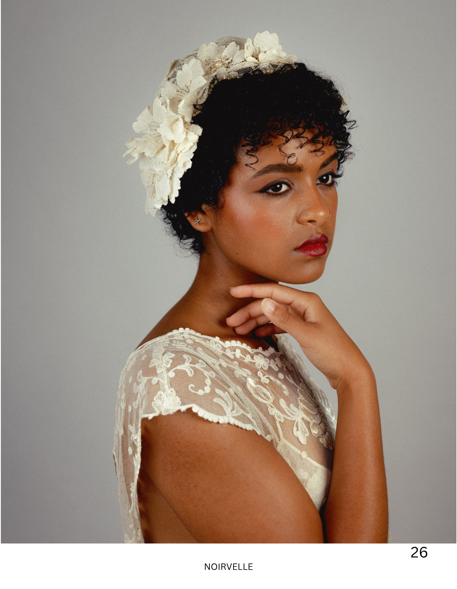 A woman with dark curly hair wearing a lace dress and a cream-colored lace floral headpiece, posing against a plain grey background.