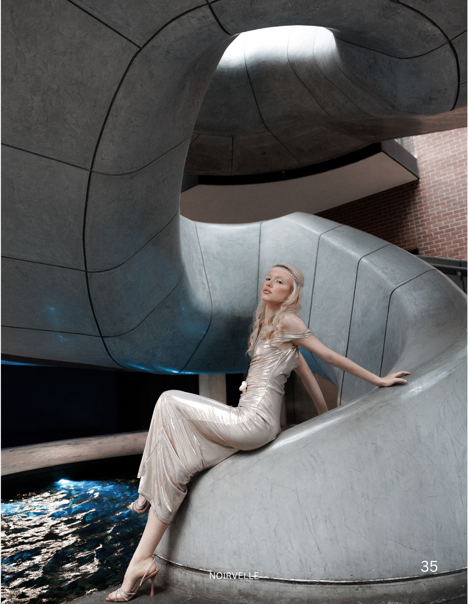 A woman in a shiny gold gown and high-heeled shoes sitting on a curved metallic structure near water. The structure has an avant-garde design with smooth, organic shapes. There is a brick building visible in the background, and the woman has long, wa