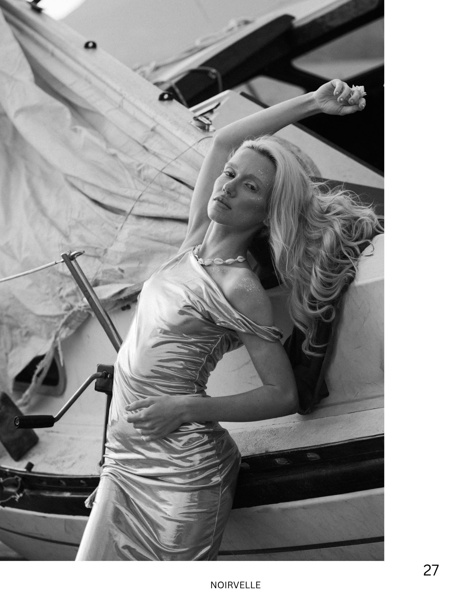 A woman with long, wavy hair lying on a boat, wearing a shiny dress and a shell necklace, with glitter on her face, holding an oar.