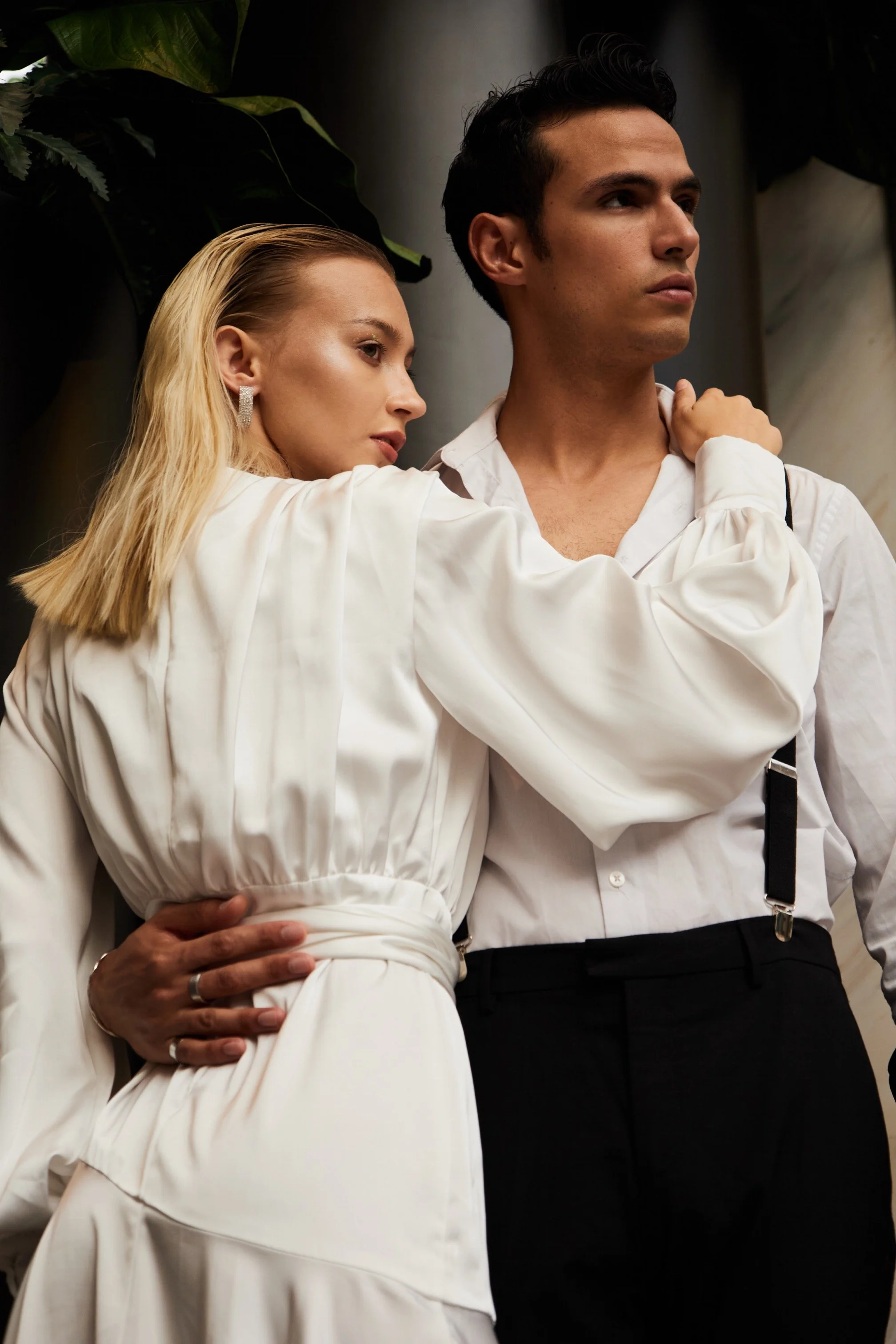 A man with dark hair and a woman with blonde hair, both dressed in white, in an intimate pose, with the woman's arm around the man's shoulder and her hand on his back, in front of a dark background with green foliage.