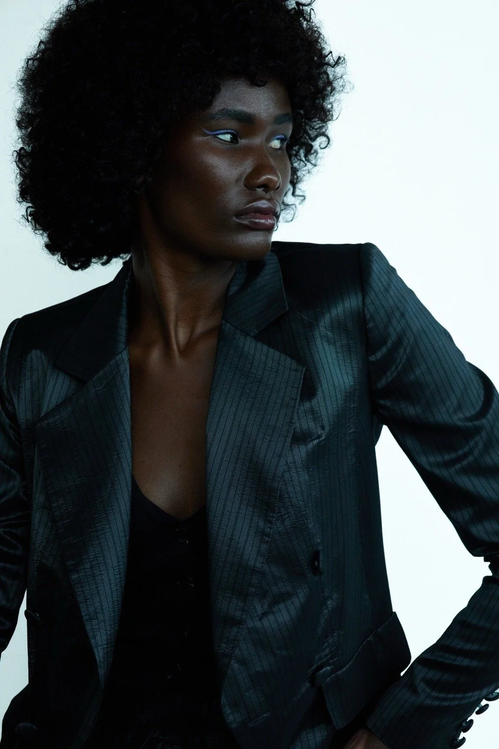 A woman with dark skin and curly black hair wearing a black pinstriped blazer and a black top, looking to the side against a white background.