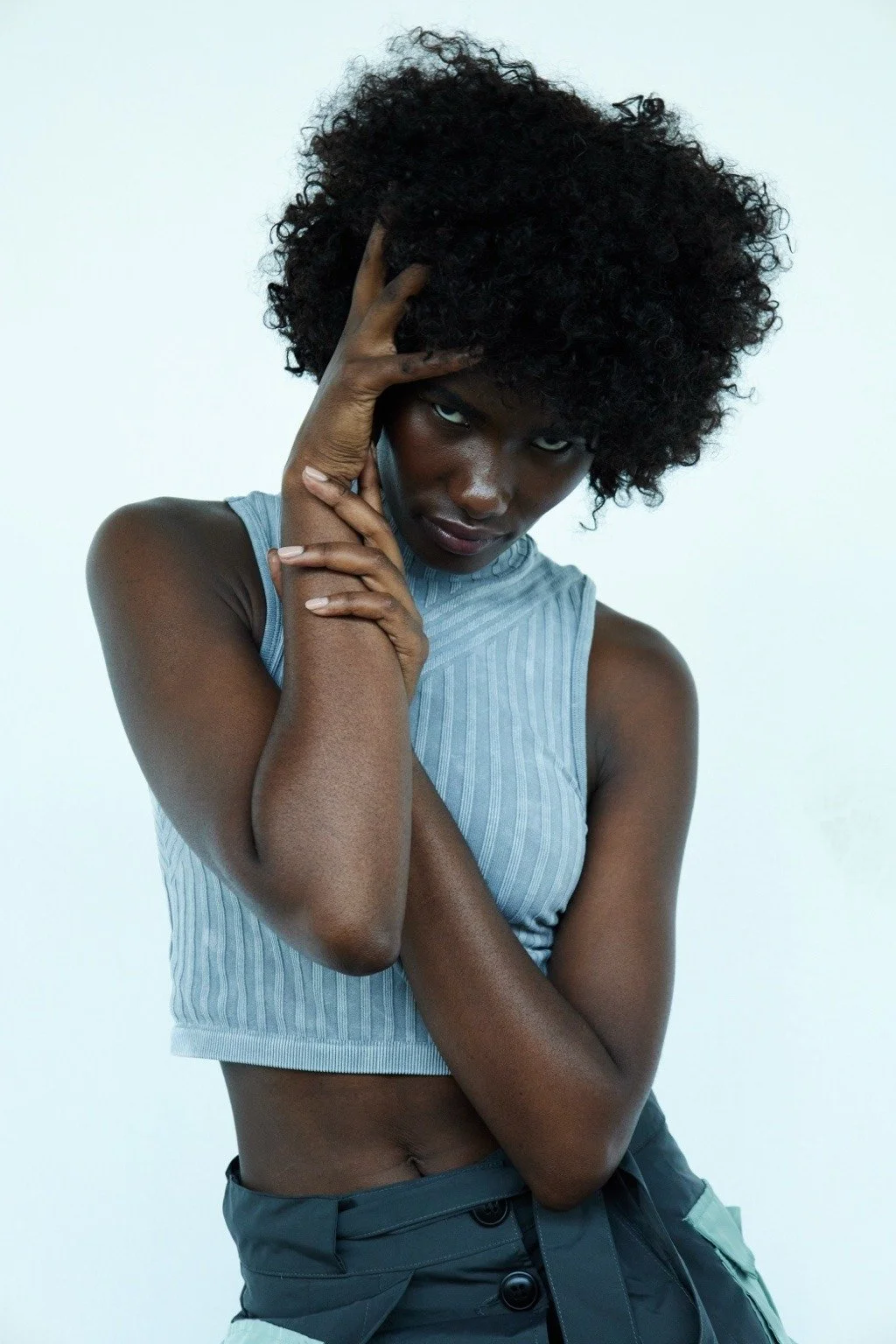 A woman with dark skin and curly black hair poses with her hand on her head and her other arm crossed, wearing a sleeveless cropped top and dark pants against a plain white background.