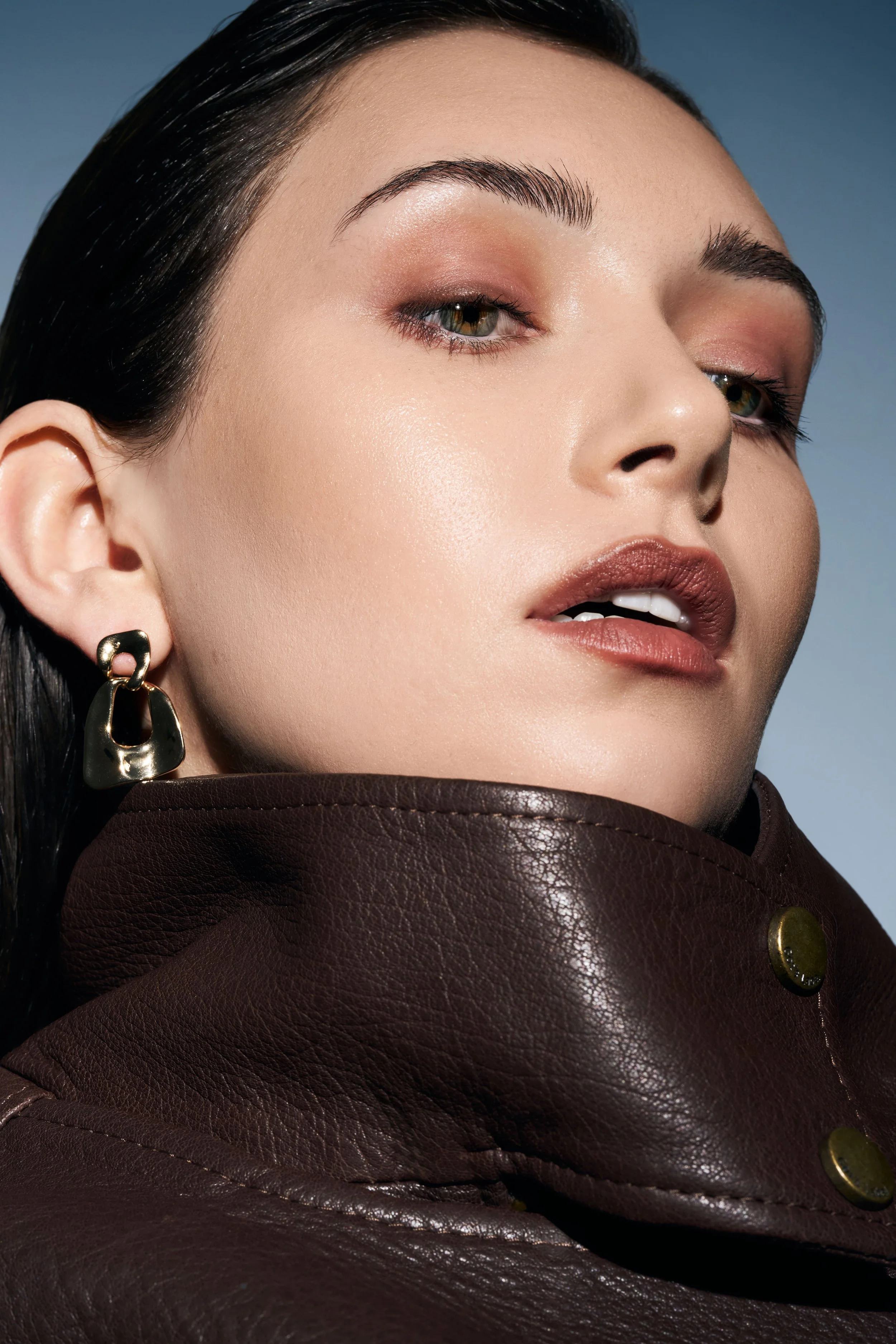 Beauty portrait of Noirvellel wearing gold earrings and a brown leather jacket against a blue background.