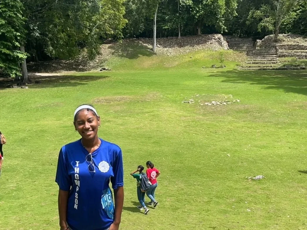 A girl smiling and wearing a blue shirt in a grassy outdoor setting with trees and ruins in the background. Two other girls are walking behind her.