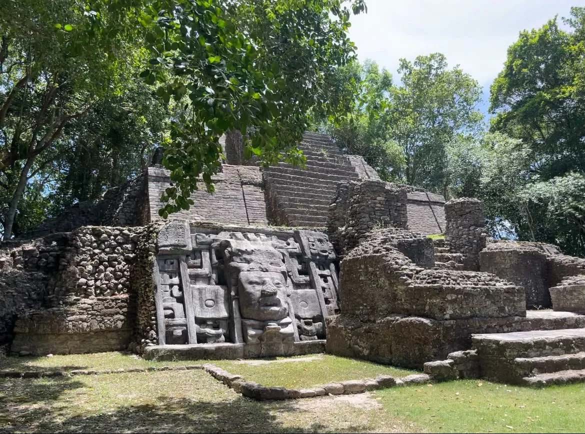 Ancient stone pyramid ruins with a carved piedra mask sculpture in the foreground, surrounded by trees and greenery.