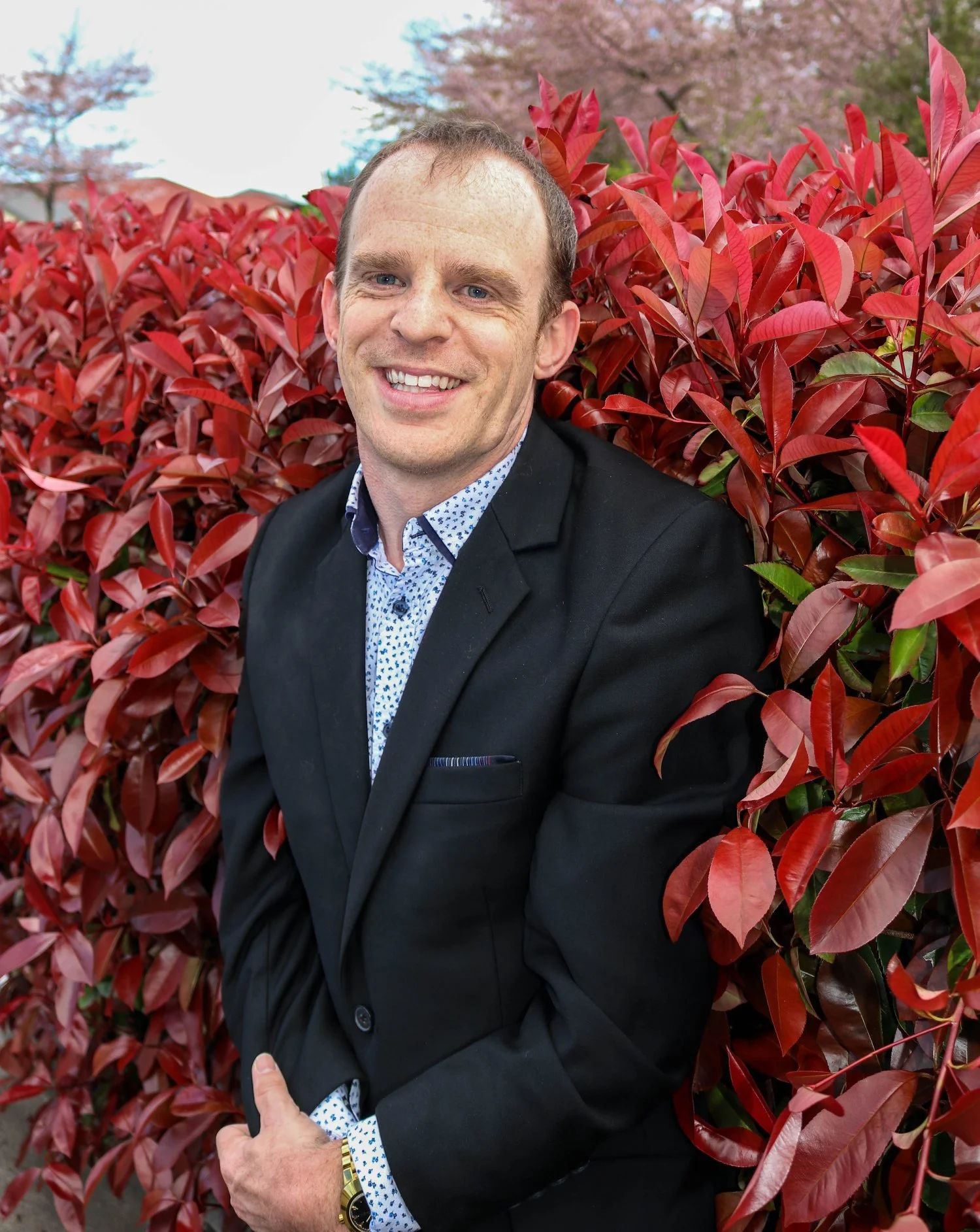 A man smiling, dressed in a black suit and white dress shirt with a small blue pattern, stands in front of vibrant red and pink leaves, with a background of a tree and partially cloudy sky.