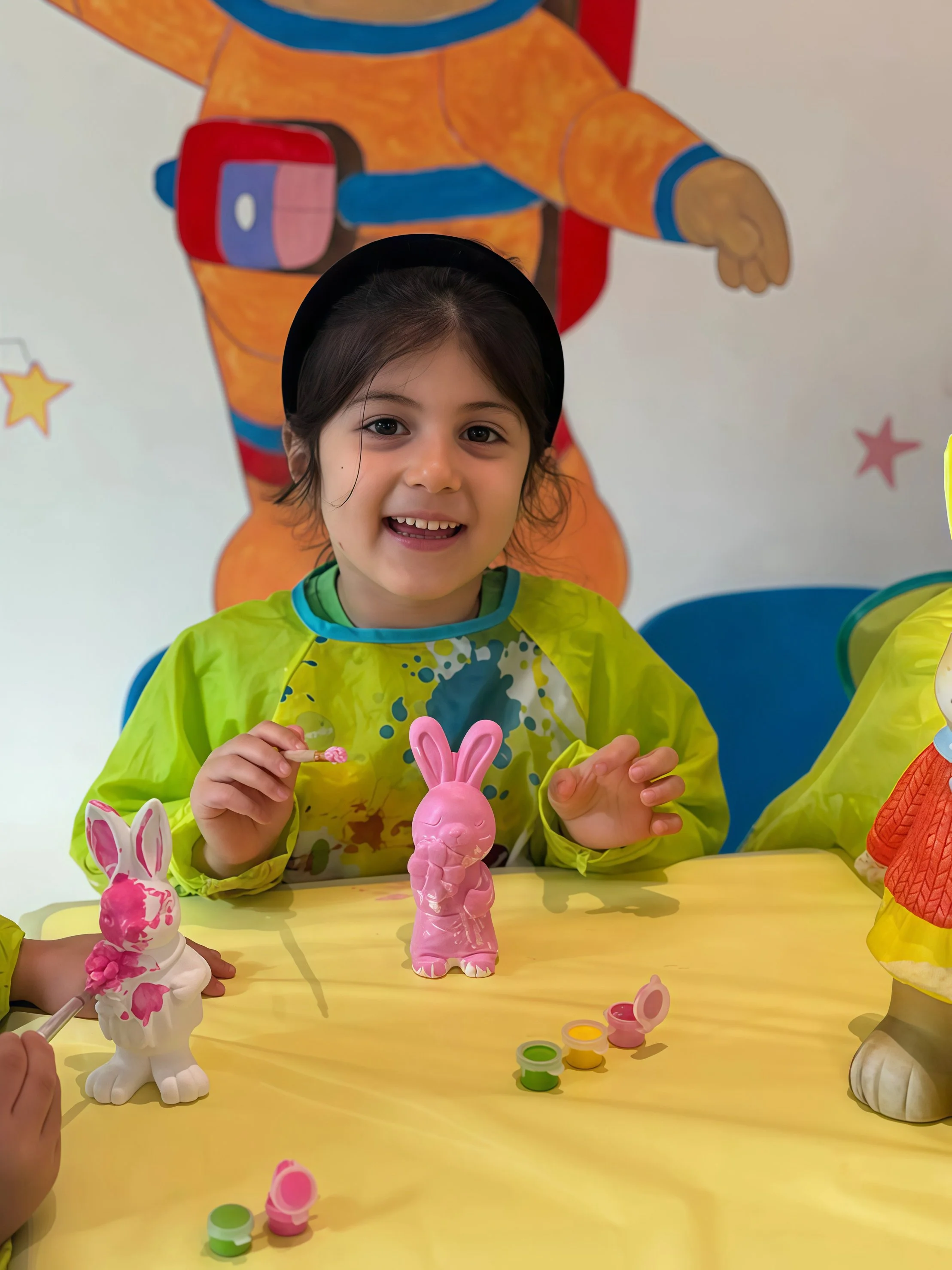 Smiling young girl with dark hair, wearing a bright green smock, painting a pink bunny figurines at a table decorated with Easter-themed toys, with colorful wall art including a cartoon carrot character in the background.