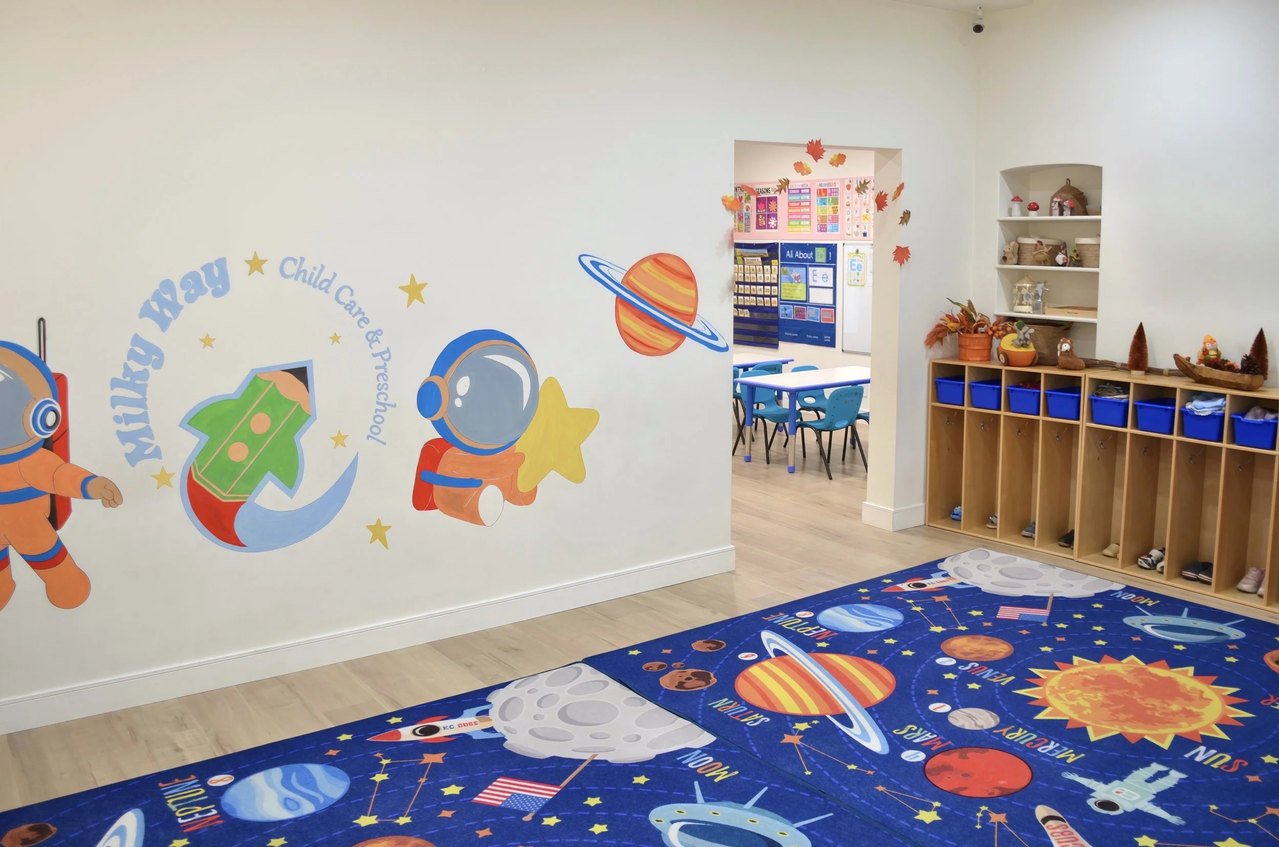 An indoor space with a space-themed design for children, featuring a wall mural with planets, stars, and an astronaut. There are tables and chairs in a classroom setting, and a wooden storage unit with blue cubbies filled with shoes or belongings. The floor has a space-themed rug and wall decorations with autumn leaves.