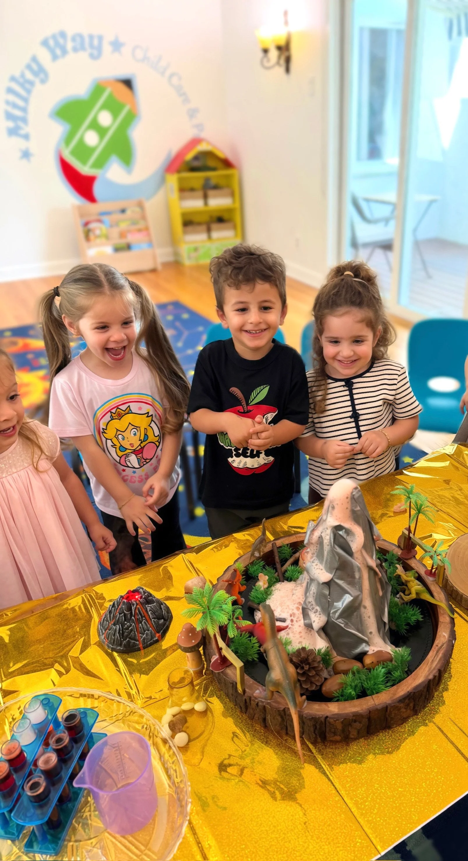 Children at a classroom gathered around a volcano science project with dinosaur-themed decorations.