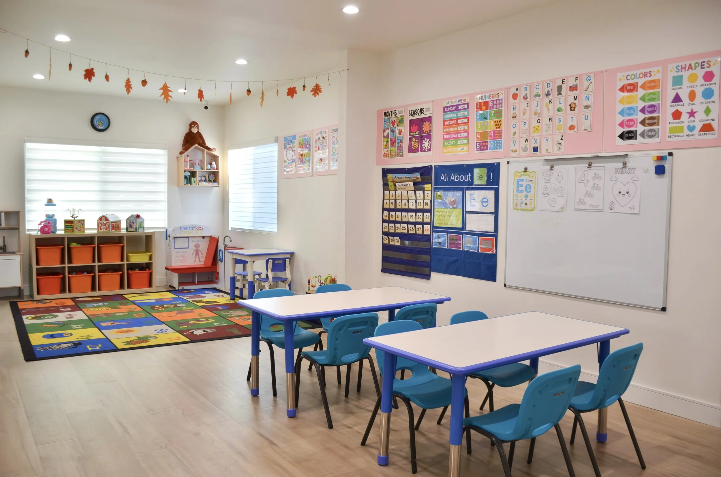 Bright, organized preschool classroom with blue chairs, a colorful alphabet rug, educational posters on the walls, and a plush toy sitting on a shelf.