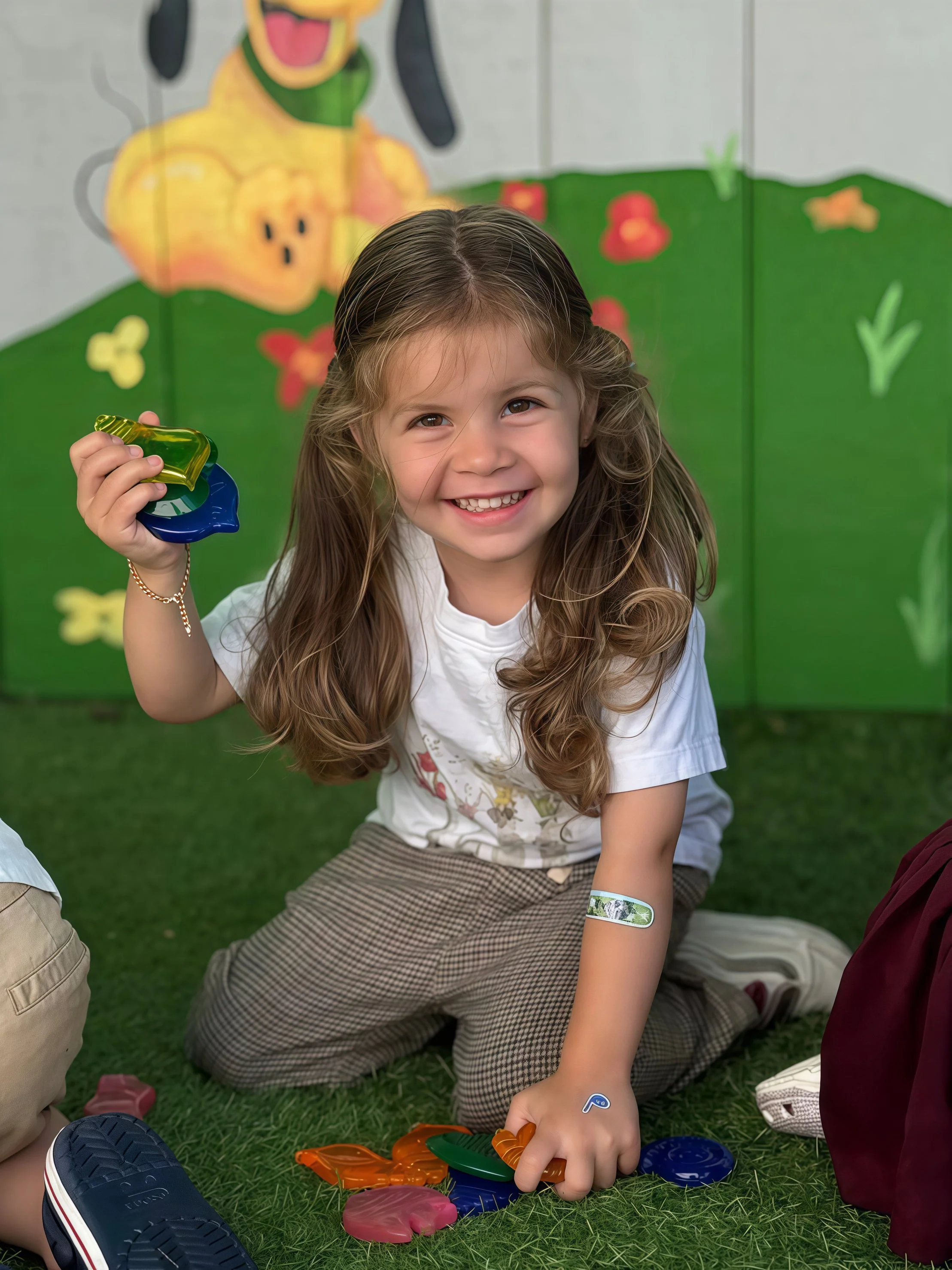 A young girl with long wavy hair, smiling and playing on green grass, holding colorful game pieces, with a painted backdrop of a cheerful dog with a yellow coat and long black ears, green collar, and a happy expression.