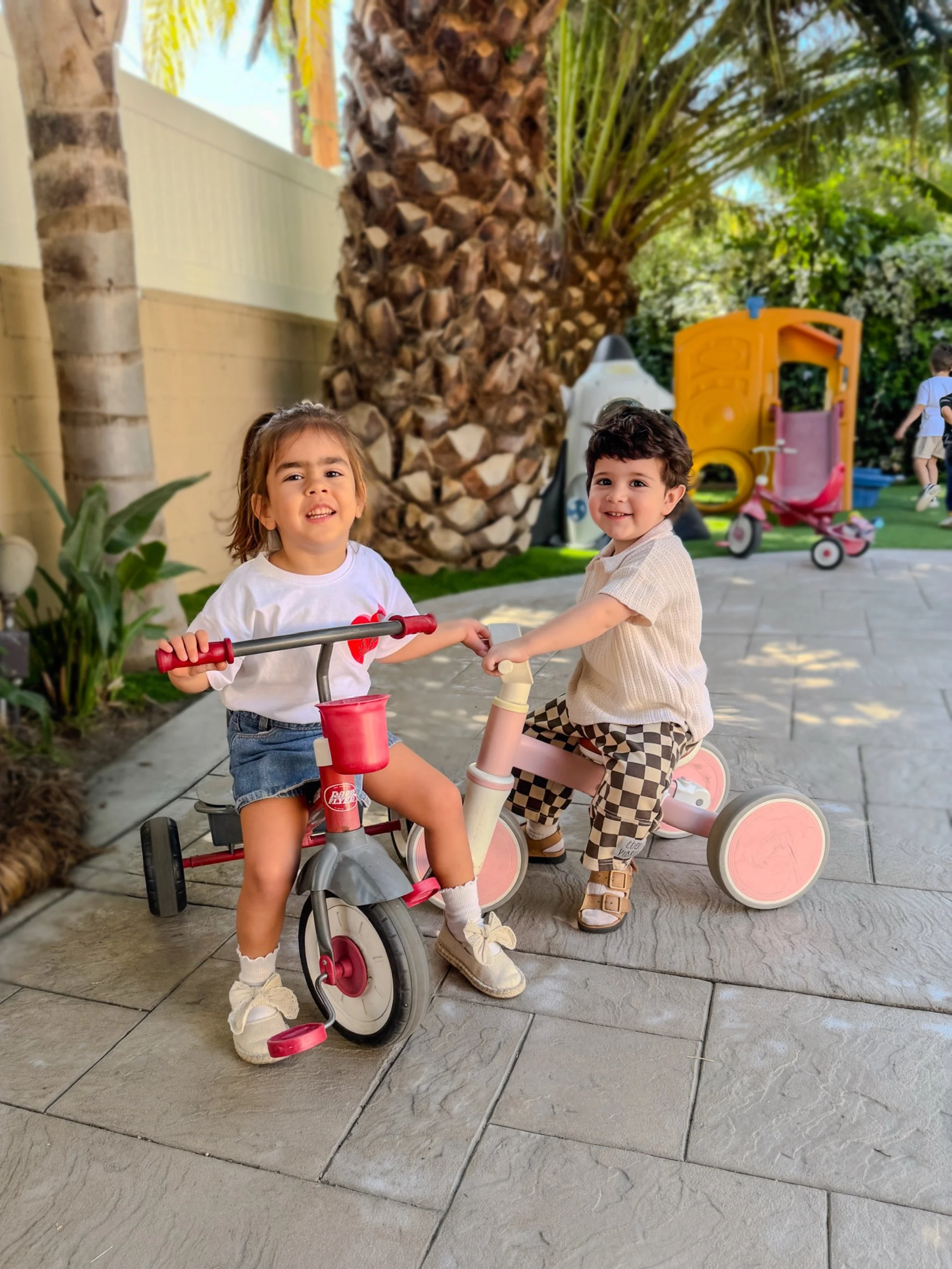 Two young children, a girl and a boy, playing together outside with tricycles, smiling and holding hands, in a backyard with palm trees, a yellow playhouse, and other children in the background.