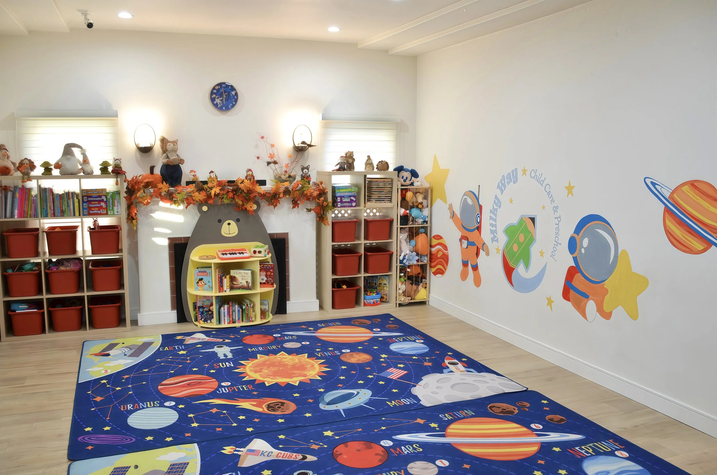 A children's classroom with space-themed decor, including a rug featuring planets and the solar system, shelves with toys and books, and a mountain-shaped bookshelf.