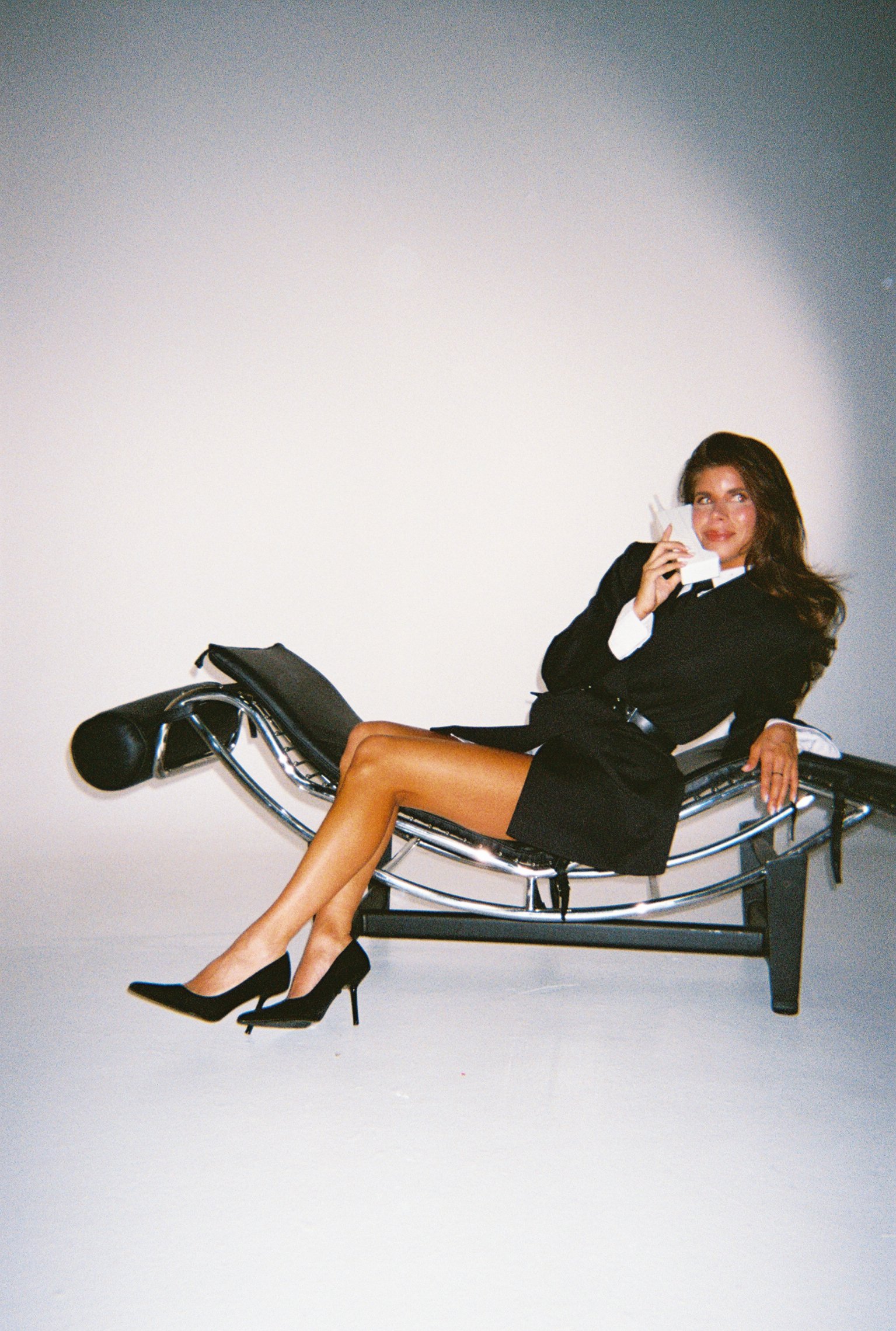 A woman dressed in black business attire sitting on a medical examination chair, holding a telephone receiver to her ear and smiling, with her legs crossed, against a plain white background.