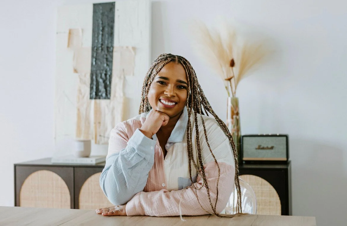 Smiling woman with braids sitting at a table in a decorated living room.