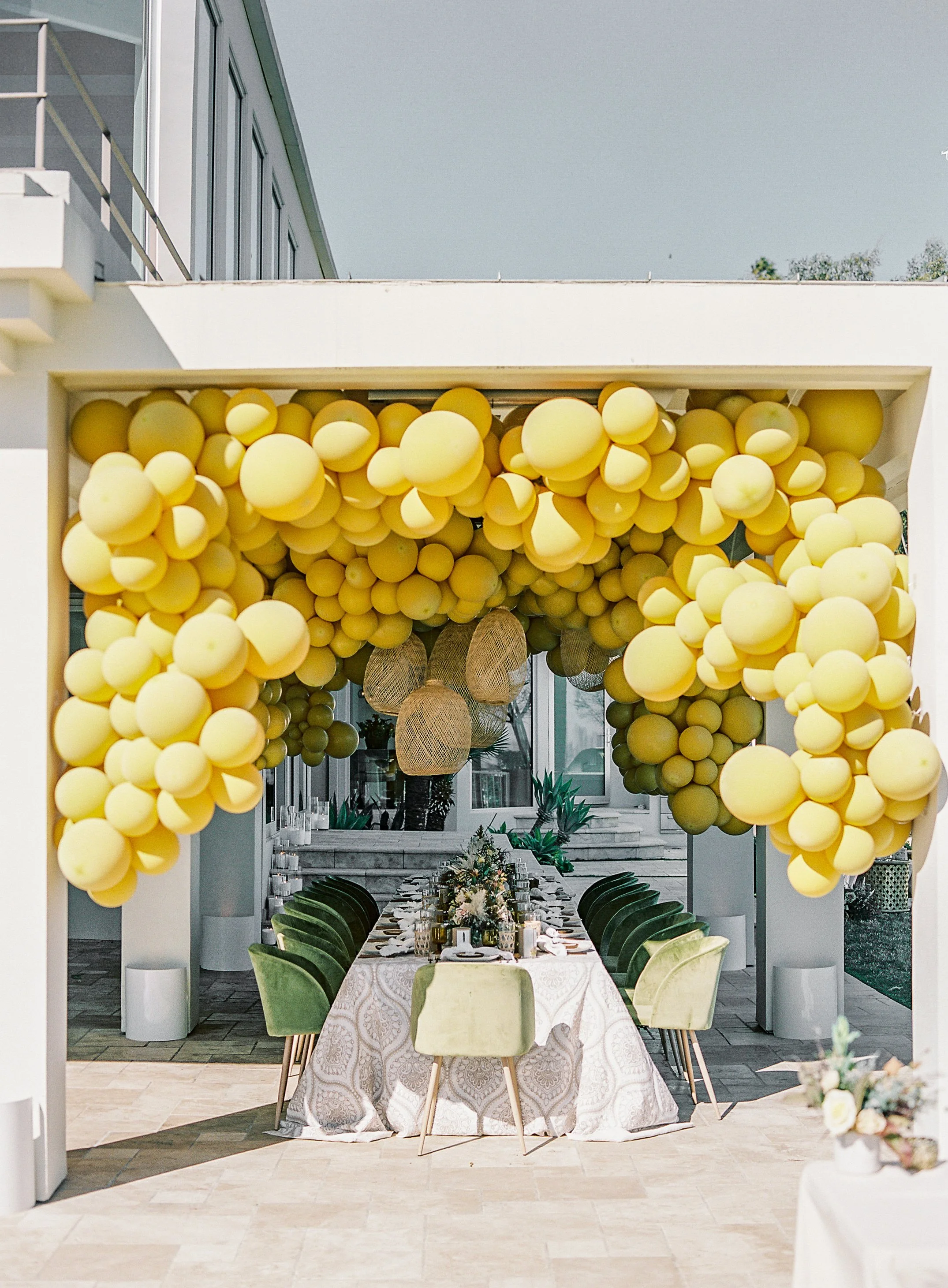 A decorated outdoor event space with a long dining table set with arrangements and chairs, adorned with a ceiling of yellow balloons and an indoor garden visible through large glass windows.