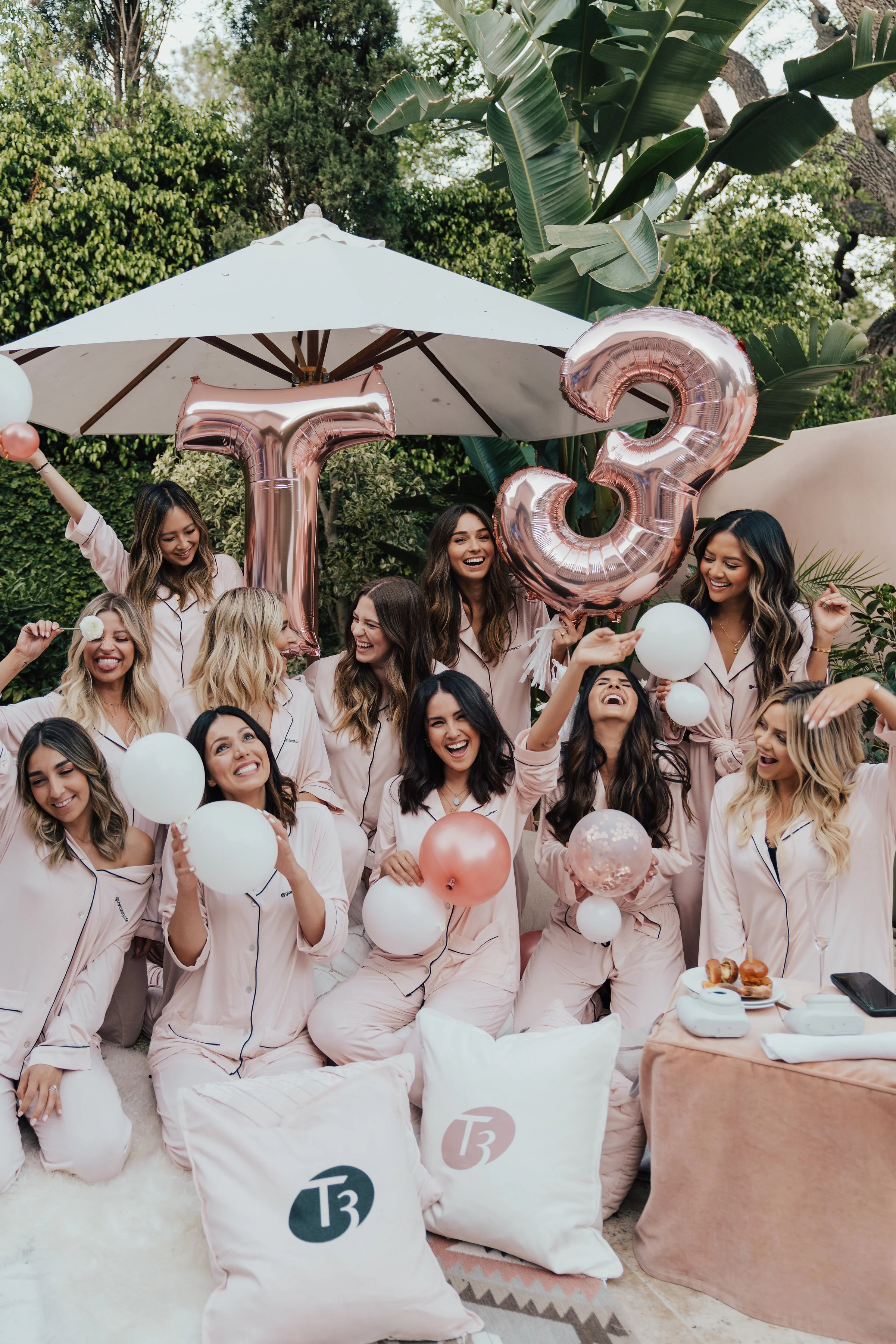 A group of women in matching pink pajamas celebrating a birthday in an outdoor garden. They are holding balloons, including large rose gold number balloons '1', '3', and '2'. There is a white umbrella, greenery, and a table with food and drinks.