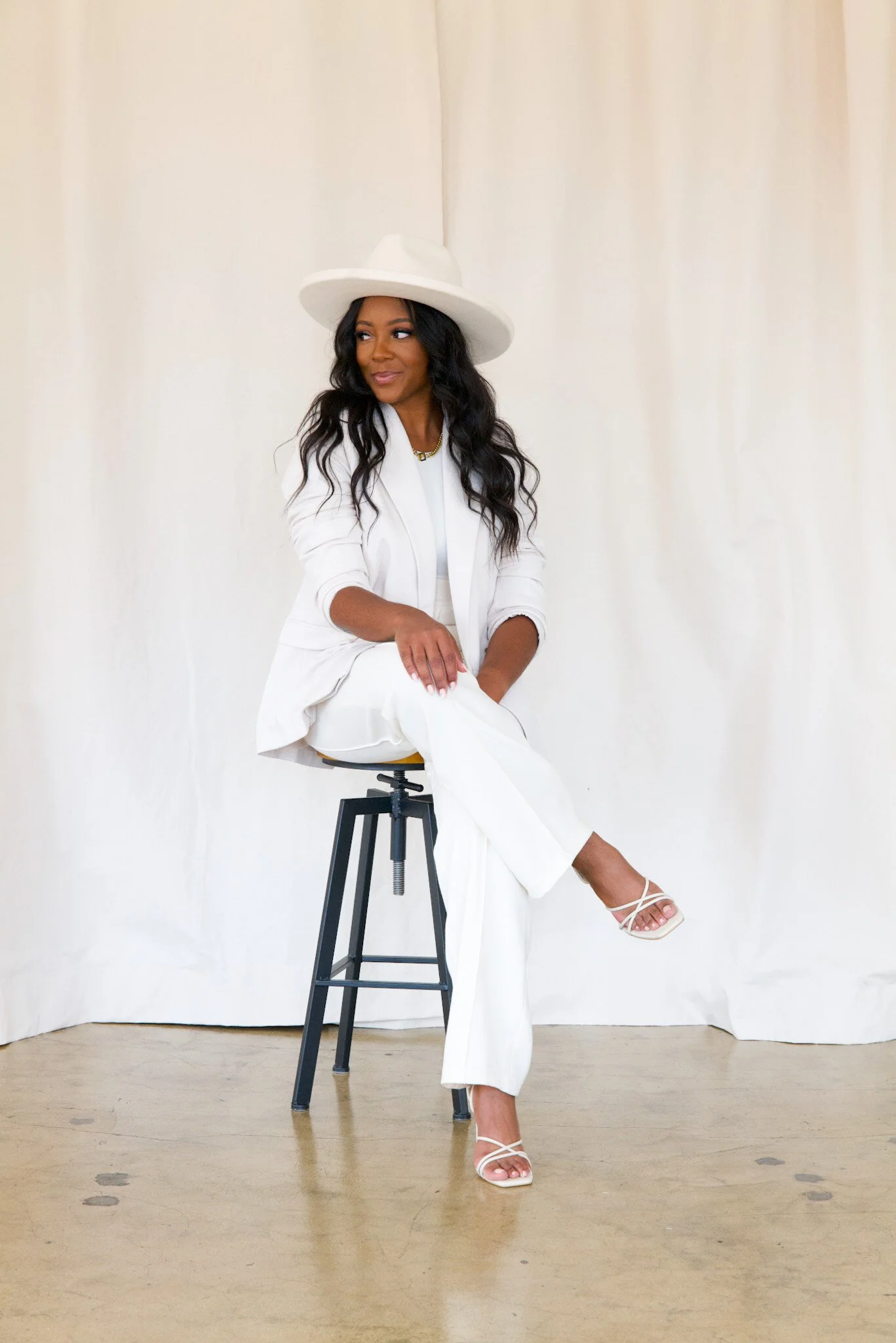 Woman sitting on a stool against a white backdrop, wearing a white wide-brimmed hat and white outfit with white high-heeled sandals.