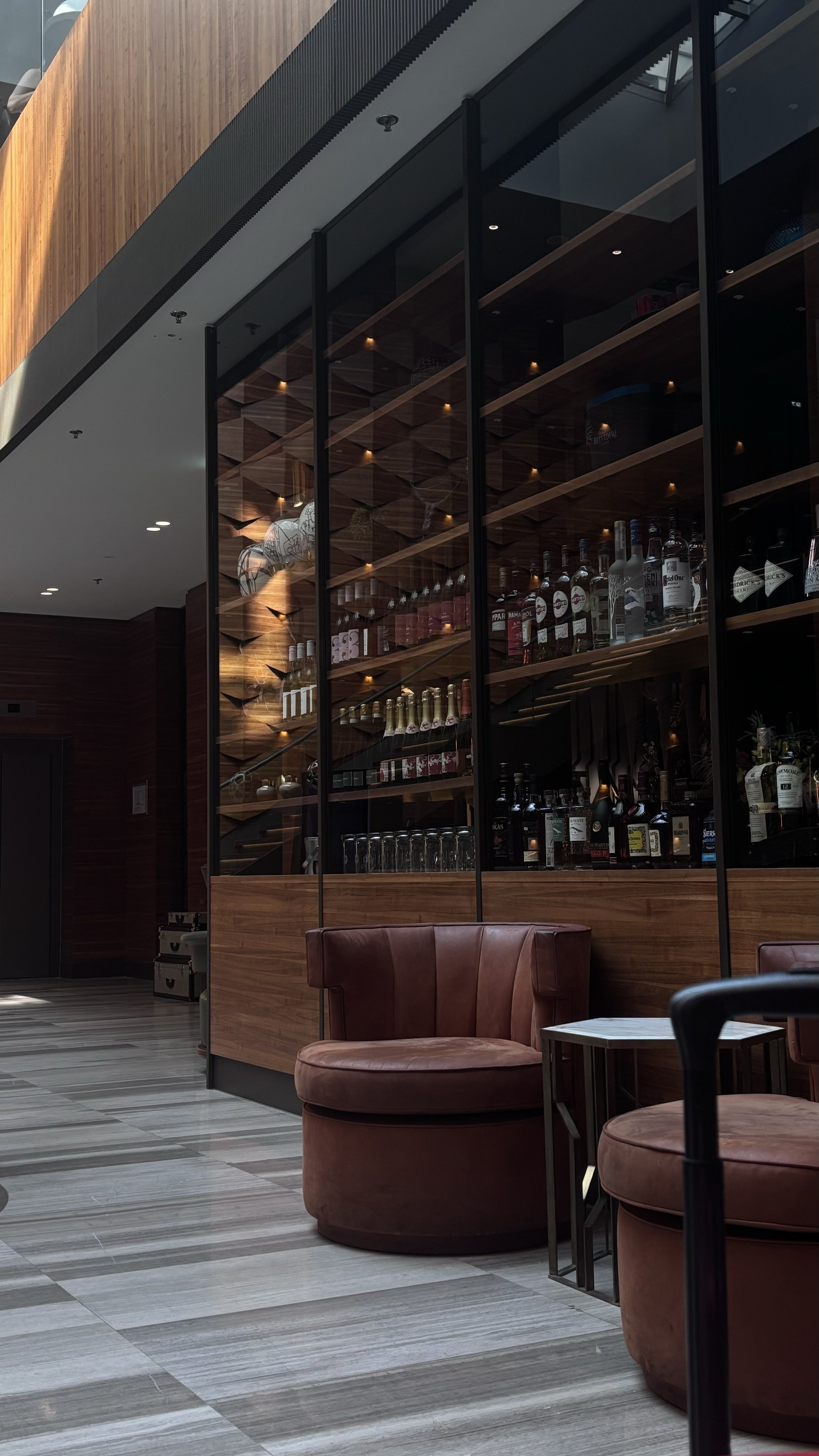 Interior of a modern hotel or office lounge with two round maroon chairs, a small side table, a glass wall with a bar behind it displaying bottles, and a wooden ceiling with small, warm lights.