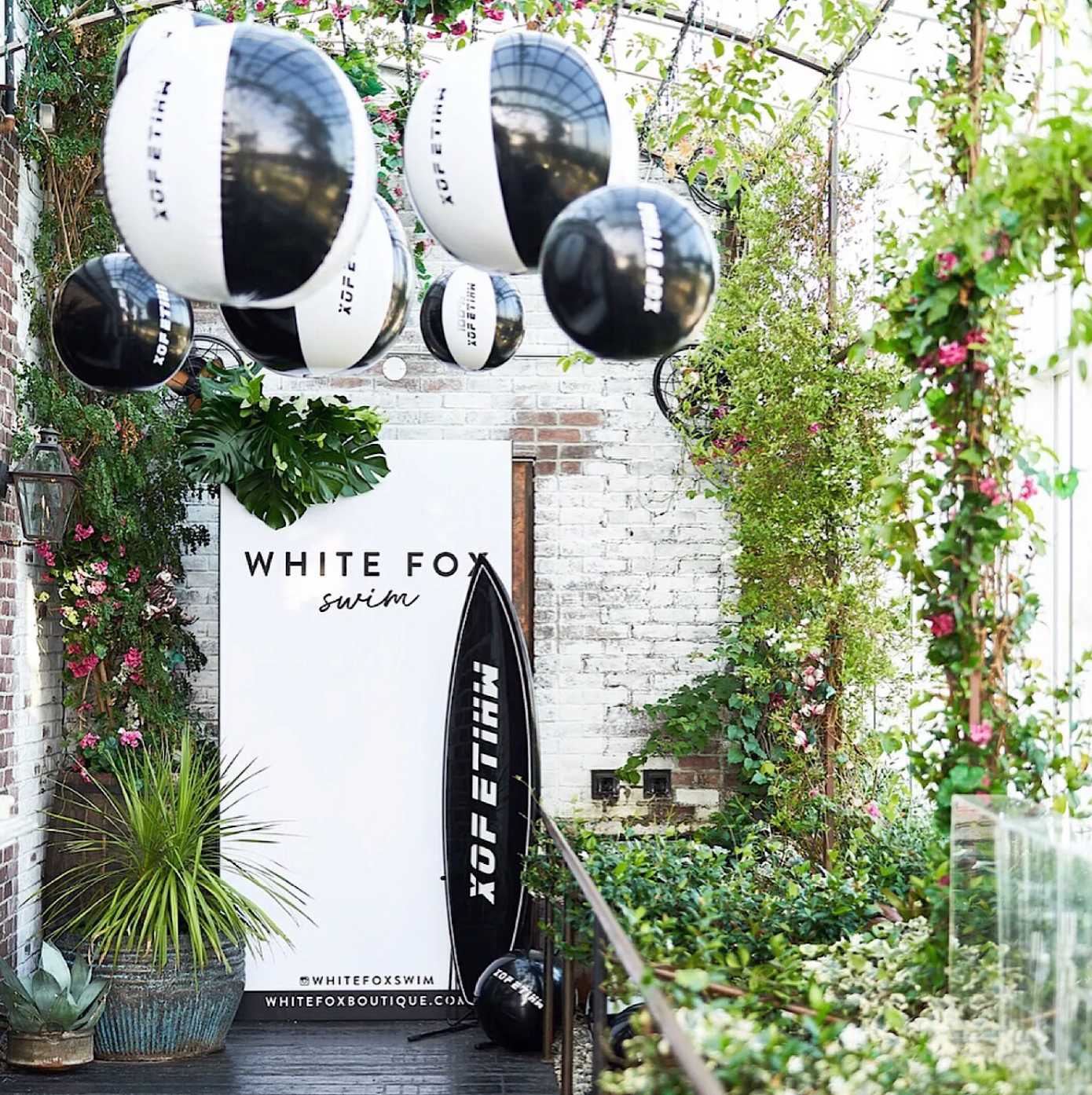 Decorative balcony with a white sign reading 'White Fox Swim', a black surfboard with the white logo, hanging black and white spherical lanterns, lush green plants, and pink flowers on a brick wall background.
