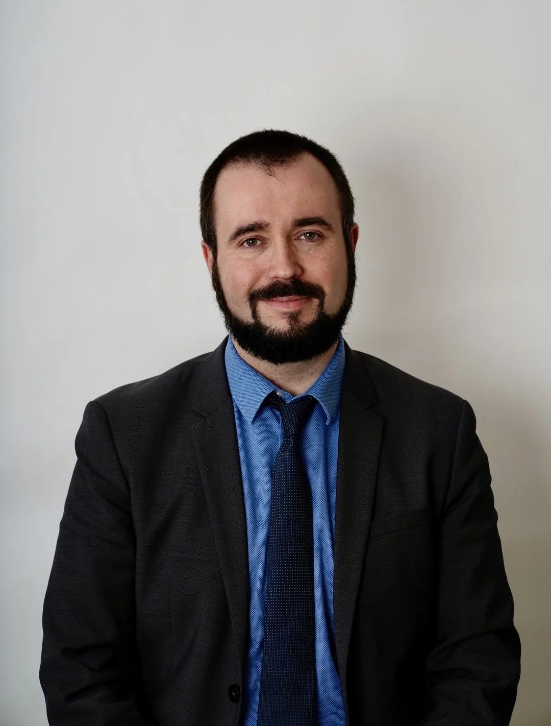 Jeune homme avec barbe, portant un costume noir, chemise bleue et cravate noire, posé contre un mur blanc.