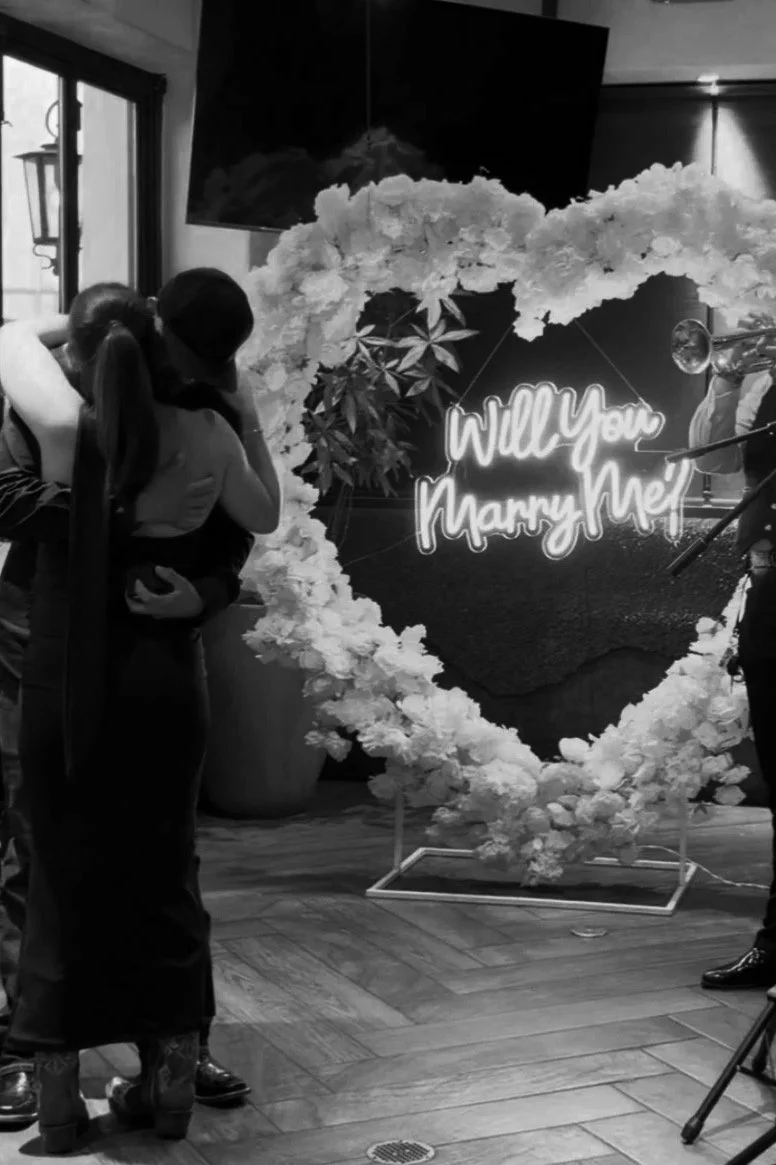 A couple sharing a kiss at a marriage proposal setup, featuring a heart-shaped floral arrangement with a neon sign that reads 'Will You Marry Me?' and a vintage gramophone in the background.