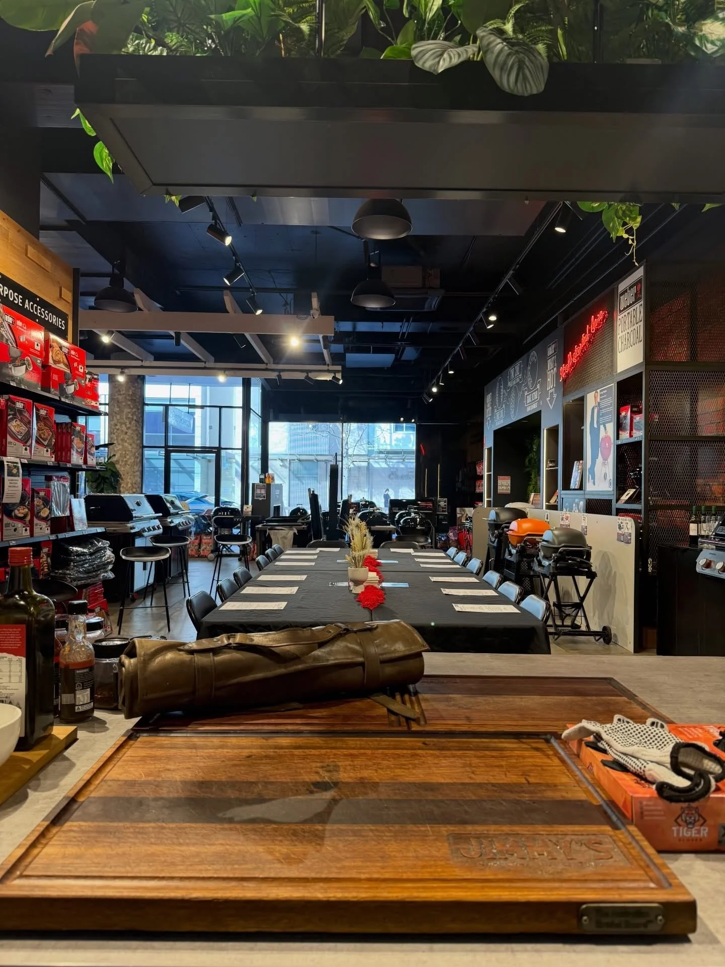 A wooden cutting board with the engraved brand logo in a restaurant or store setting, with various condiments, a leather bag, and a box of grilling tools on top, with tables and store displays in the background. In store Weber BBQ masterclass