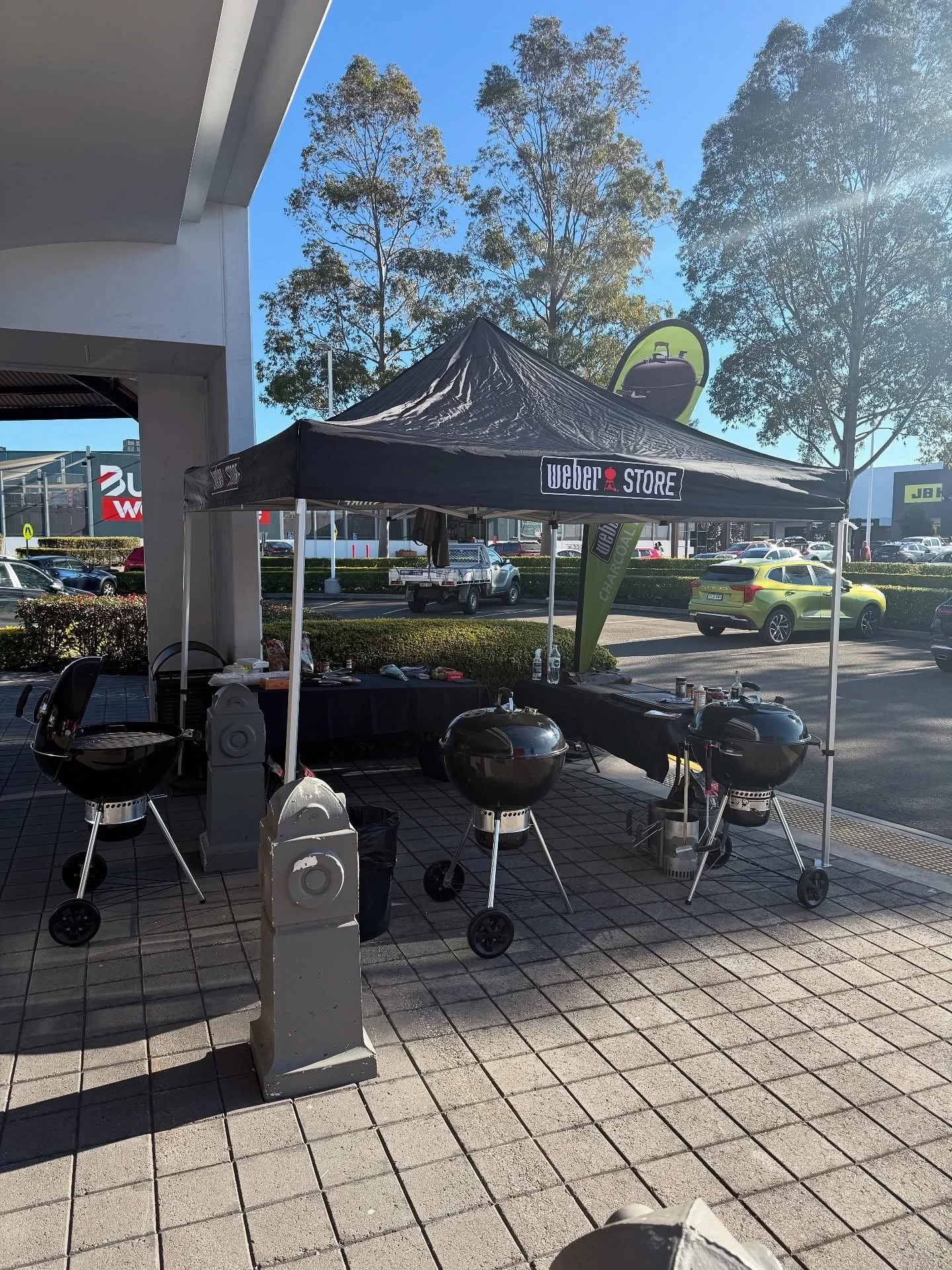 Outdoor Weber barbecue setup with multiple grills under a canopy, in a parking lot area with cars, trees, and shopping center buildings in the background. BBQ Demo