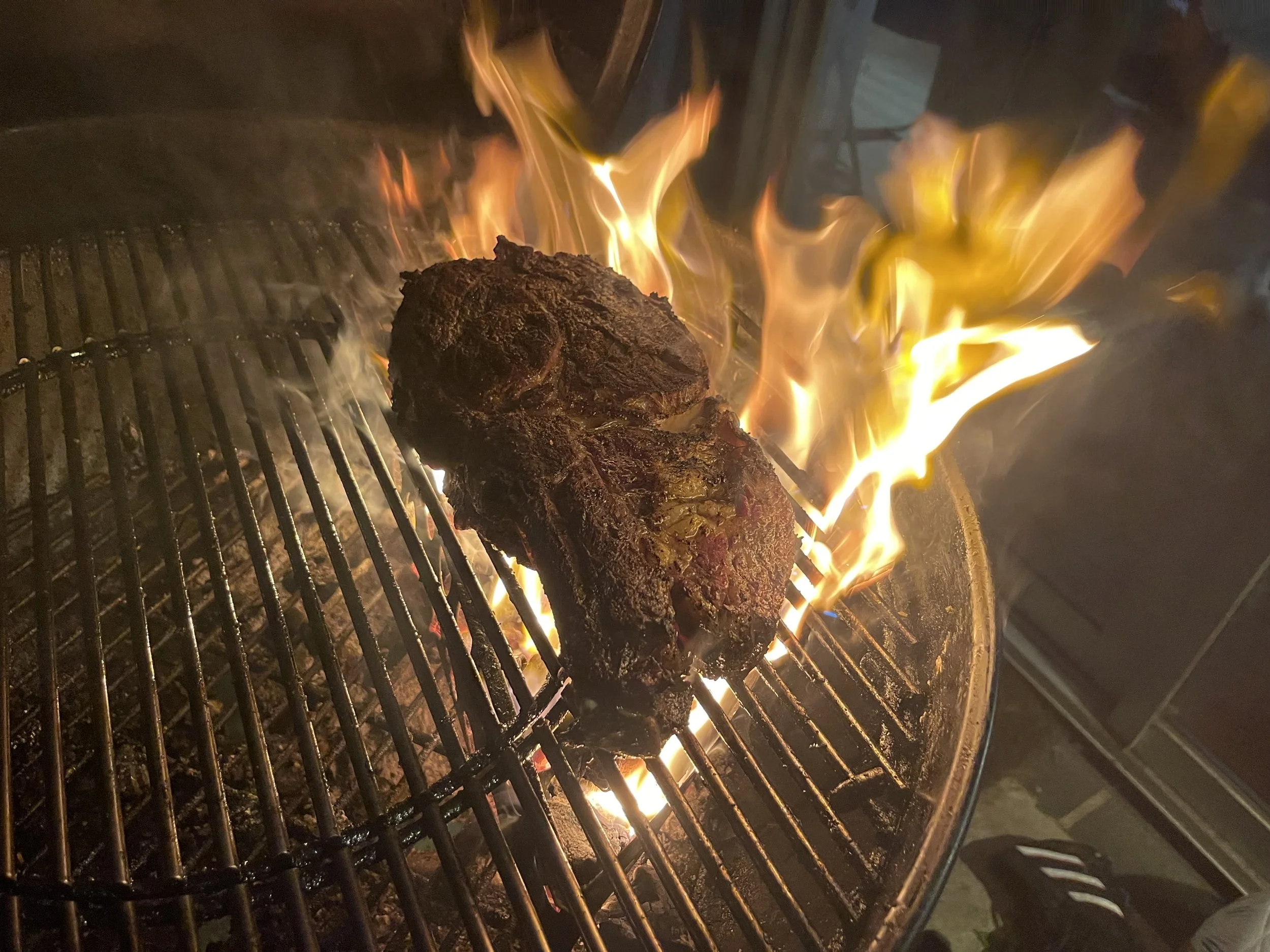 A piece of meat cooking on a barbecue grill with flames and smoke visible, grill grates underneath the meat.