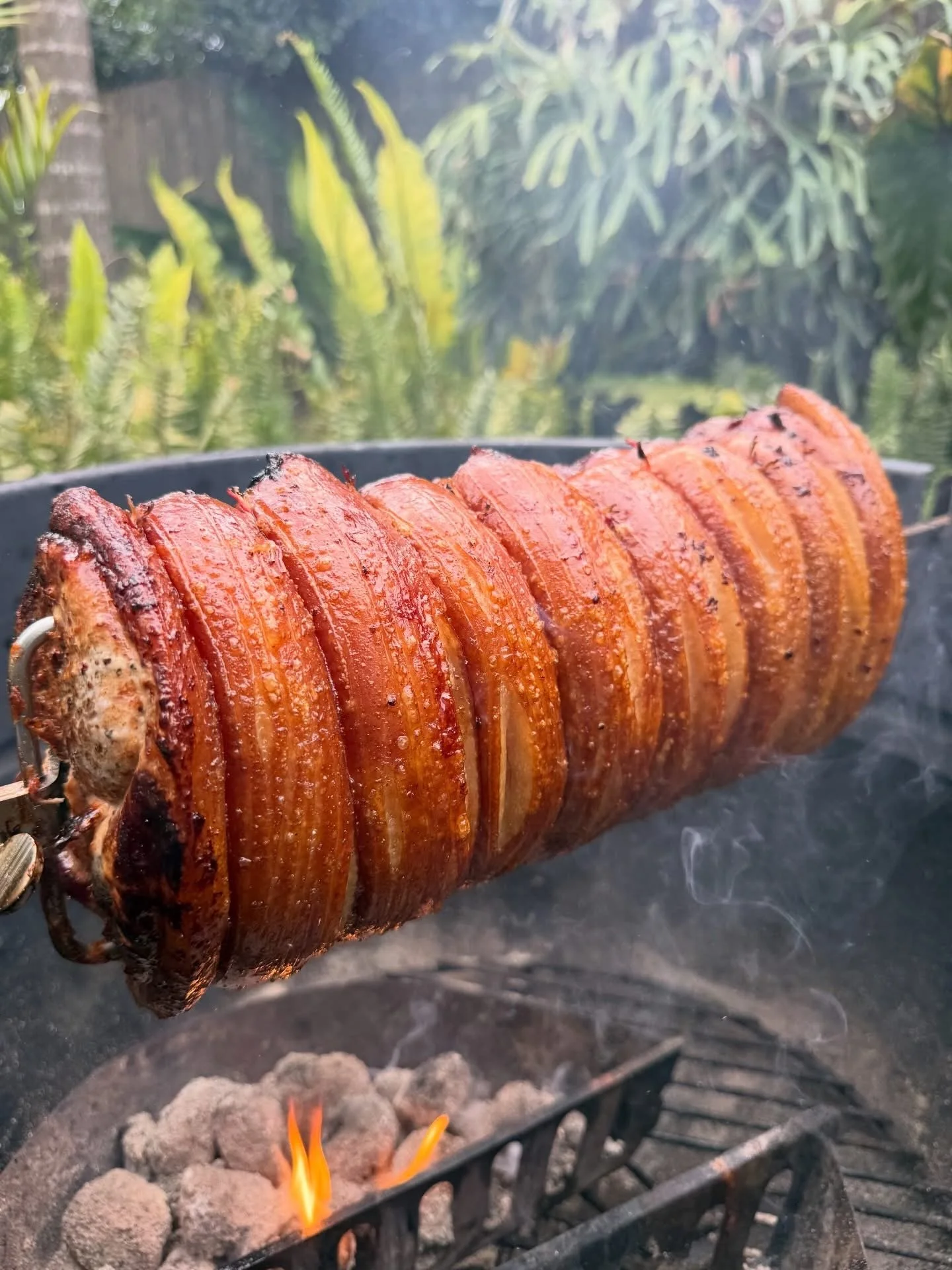 A whole pork roast on a spit roasting over an open flame grill outdoors in a garden with green plants in the background.