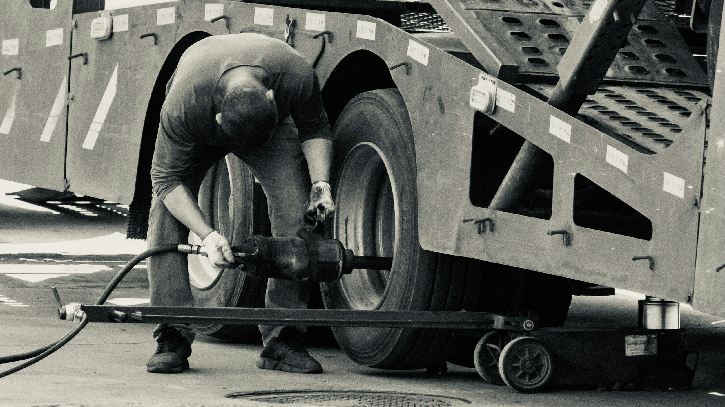 A worker in work gloves is crouched down and using a power tool to work on a large vehicle tire, with a metal frame surrounding the area.