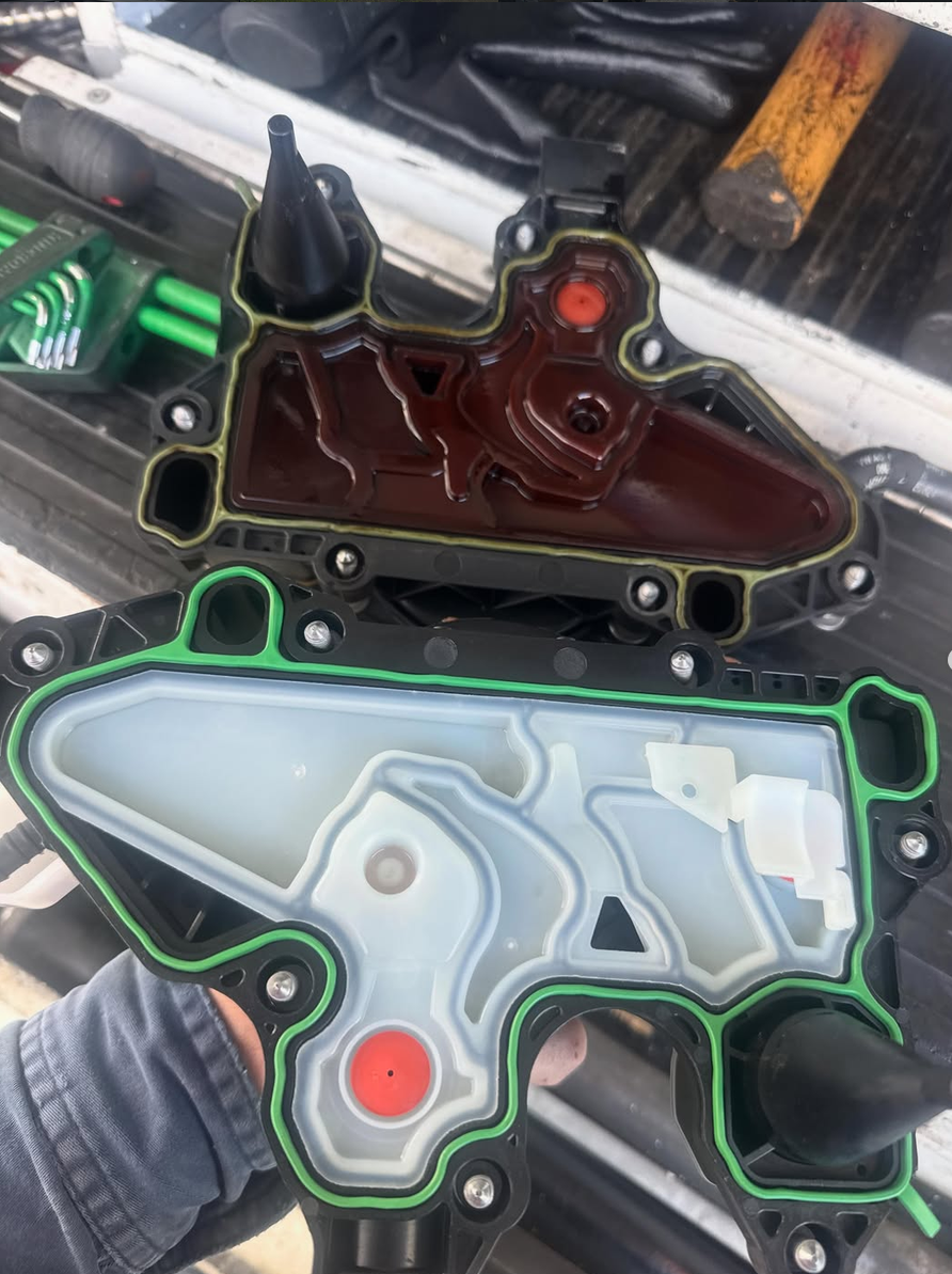 A mechanic's hand holding an automotive valve body with a gasket installed, with another valve body in the background. The valve body appears to be part of a transmission system, with various solenoids and channels visible inside.