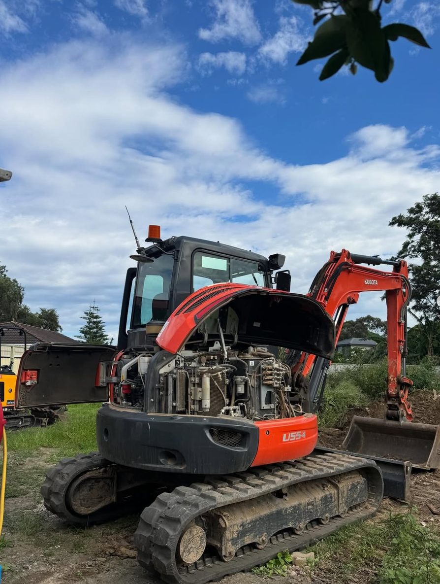 A red and black Kubota mini excavator with its engine cover open, construction site outdoors with blue sky and clouds.