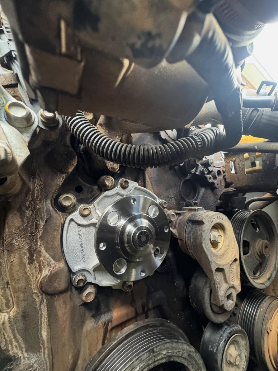 Close-up of a car engine with a new metal pulley installed near the center, surrounded by various engine parts, belts, and hoses.