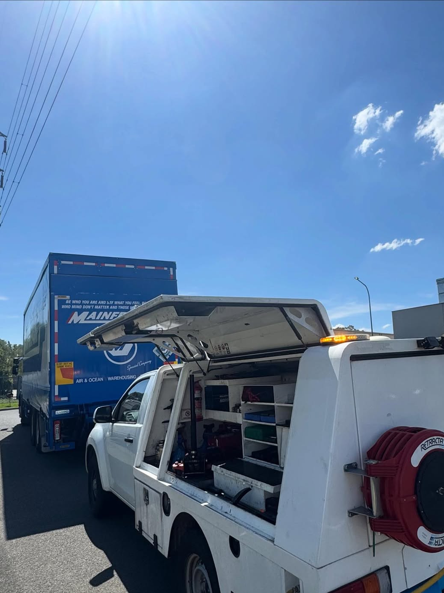 A utility truck with open compartments and equipment, parked behind a large moving truck on a sunny day with blue sky and a few clouds.