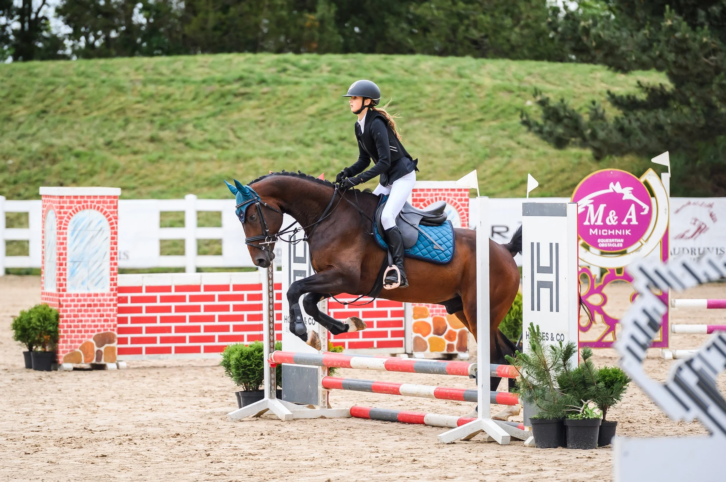 A female equestrian wearing a black helmet, black riding jacket, and white riding pants riding a horse and jumping over a show jump obstacle at an outdoor equestrian event.