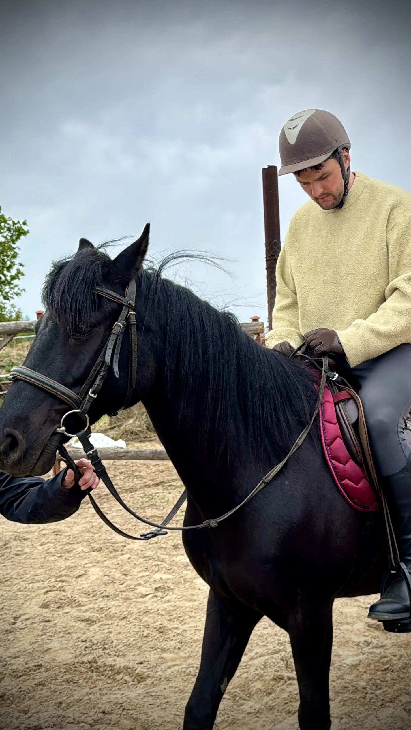 A man wearing a beige fleece jacket and riding a black horse with a red saddle pad, wearing a brown riding helmet. The man is looking down, and another person is holding the horse's bridle.