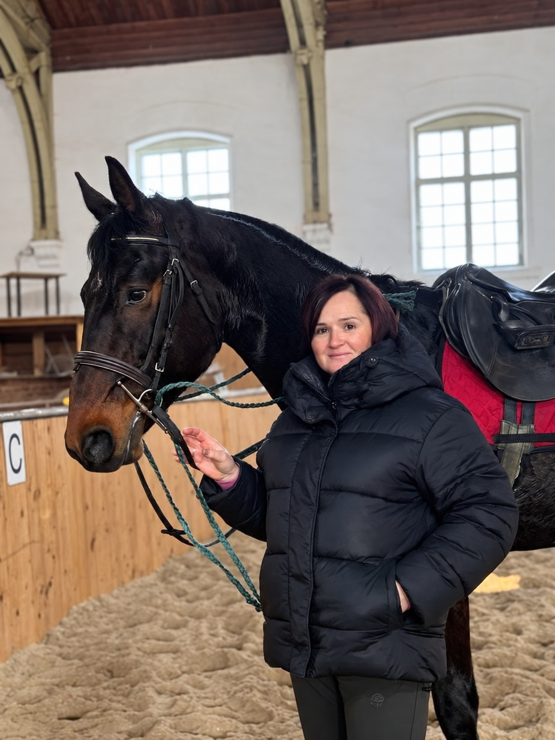 A woman standing next to a black horse inside an indoor riding arena. The woman is wearing a black puffer jacket and has short dark hair. The horse is saddled with a red saddle blanket and a black saddle.