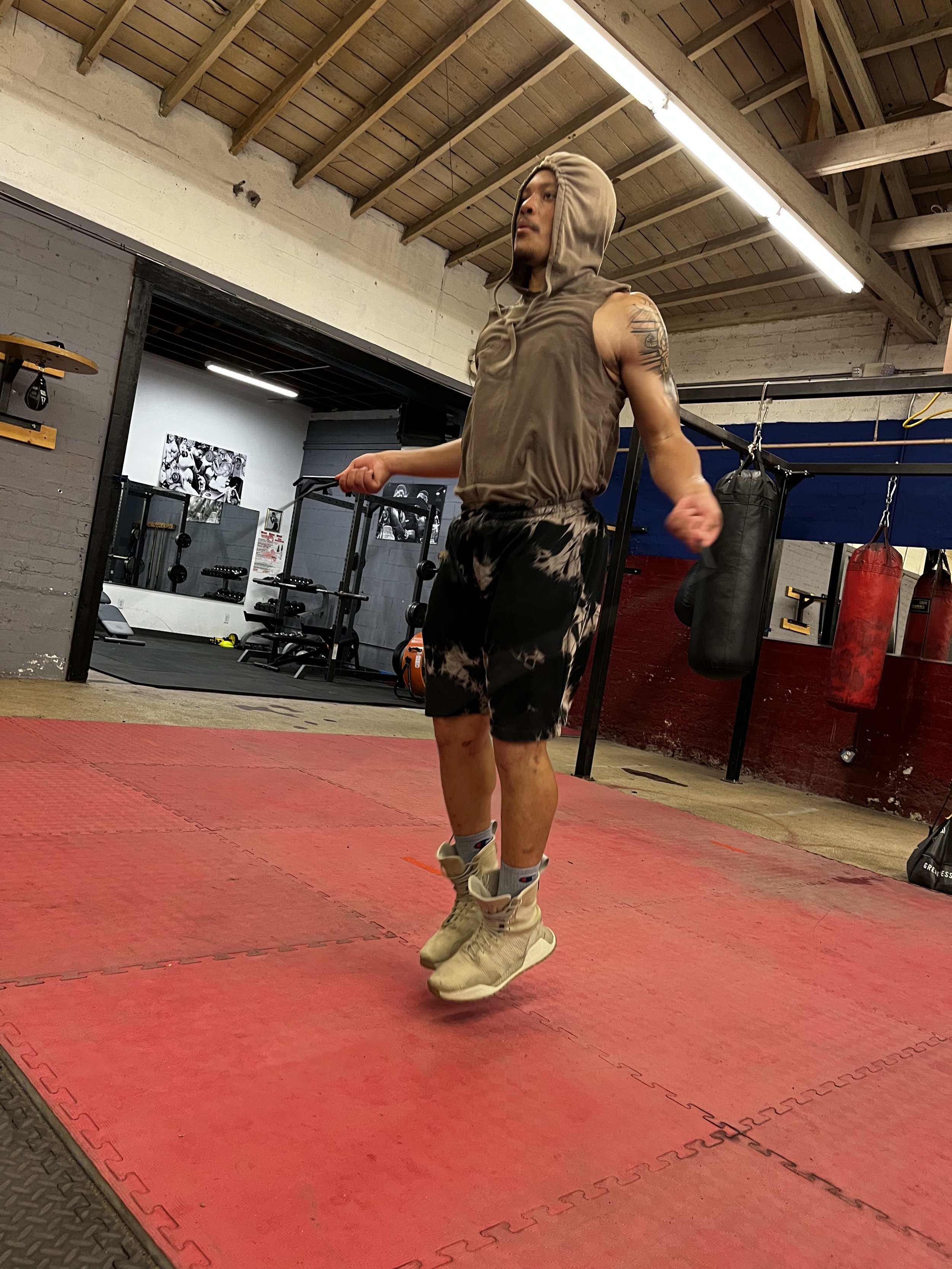 A man with a beige hoodie and patterned shorts is jumping in a gym. He is wearing beige sneakers and has tattoos on his upper arm. The gym has boxing punching bags and workout equipment in the background.