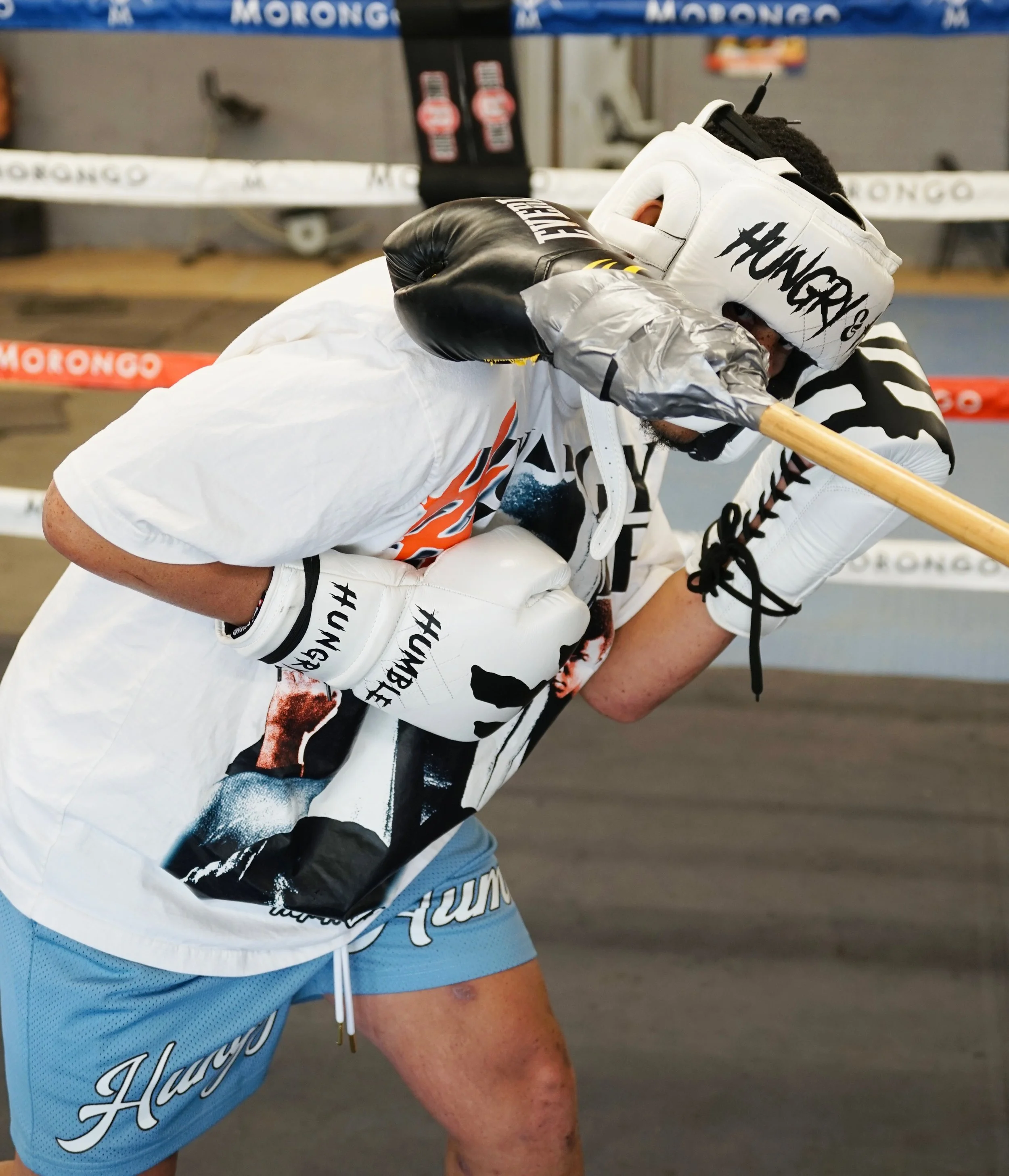 A boxer is resting with his head down and covered by a white headgear that has 'HUNG R' written on it, in a boxing gym with a punching bag in the background. He is wearing a white shirt with black and orange graphics and light blue shorts.