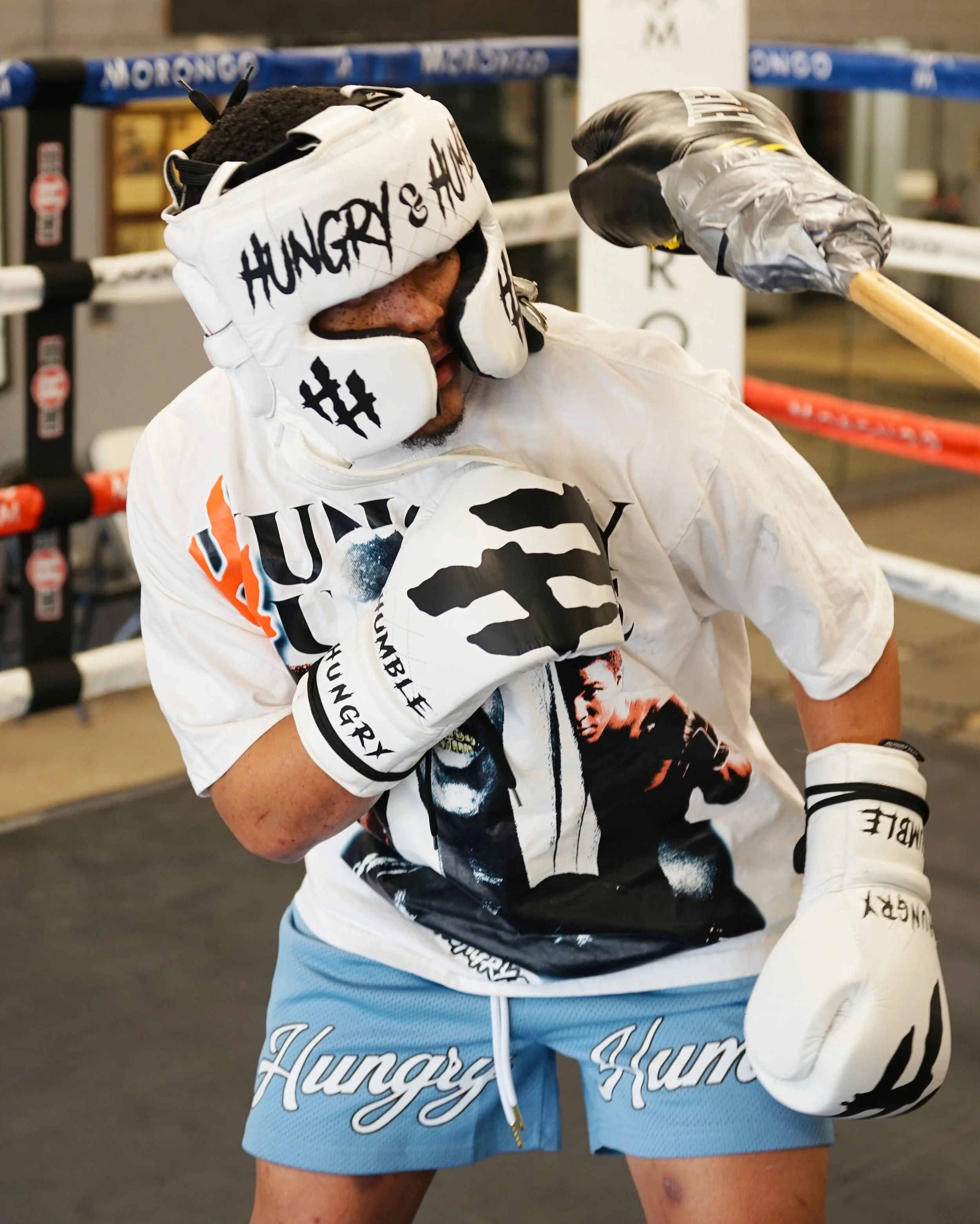 Close-up of a boxer’s face and upper body in a boxing gym. The boxer is wearing white and black gloves, a white shirt with black and orange details, and light blue shorts with the word 'Hurricane' printed on them. The boxer is throwing a punch with their glove near their face. The gym background includes boxing rings and equipment.