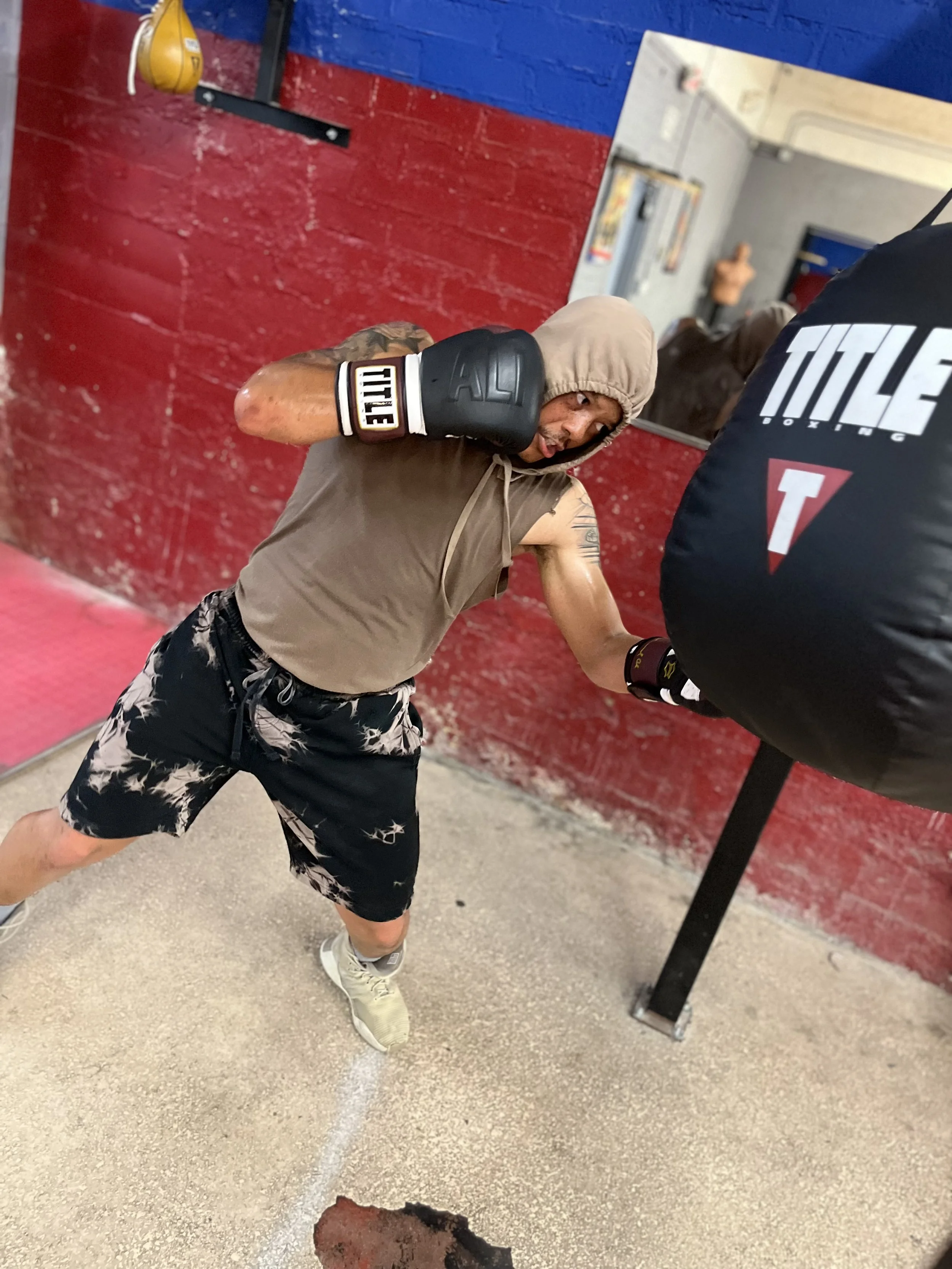 A man wearing beige shorts, a sleeveless shirt, and boxing gloves is practicing boxing with a black punching bag in a gym. The background features a red brick wall and a mirror.