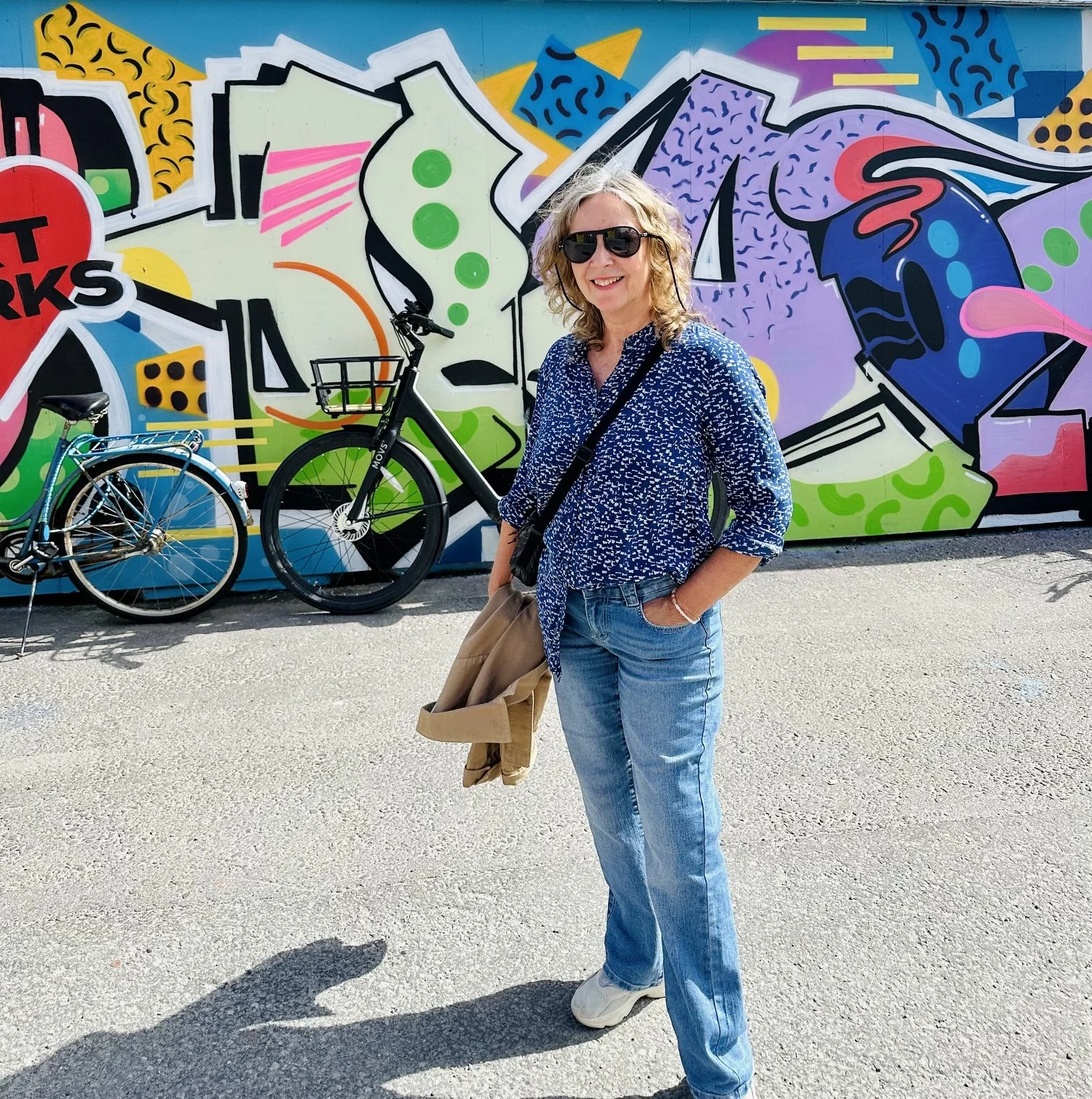 A woman with blonde curly hair wearing sunglasses, a blue patterned shirt, and jeans, standing in front of a colorful graffiti mural with two bicycles nearby.
