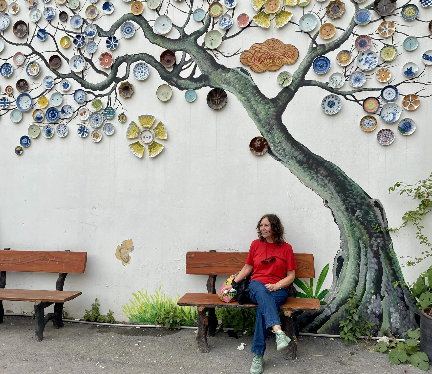 A woman sitting on a wooden park bench beneath a painted mural of a tree with decorative plates as leaves on the wall behind her. The woman is wearing a red shirt and blue jeans, with a colorful bag on her lap. There are two more benches to her left and some greenery at the base of the wall.