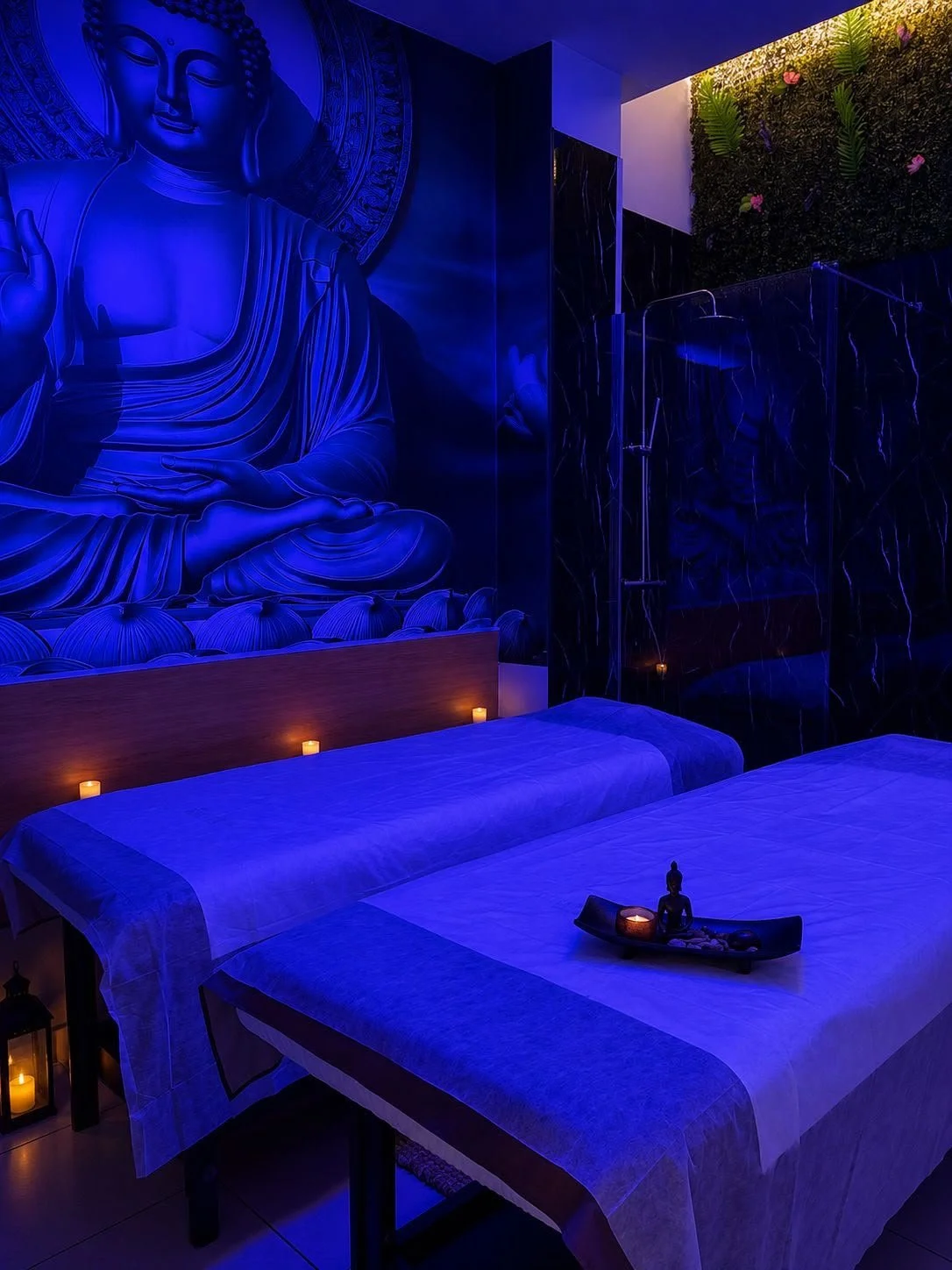 Habitación de spa con fondo de pared con estatua de Buda iluminada en azul, camas con blancos, vela y decoración zen en un ambiente relajante.