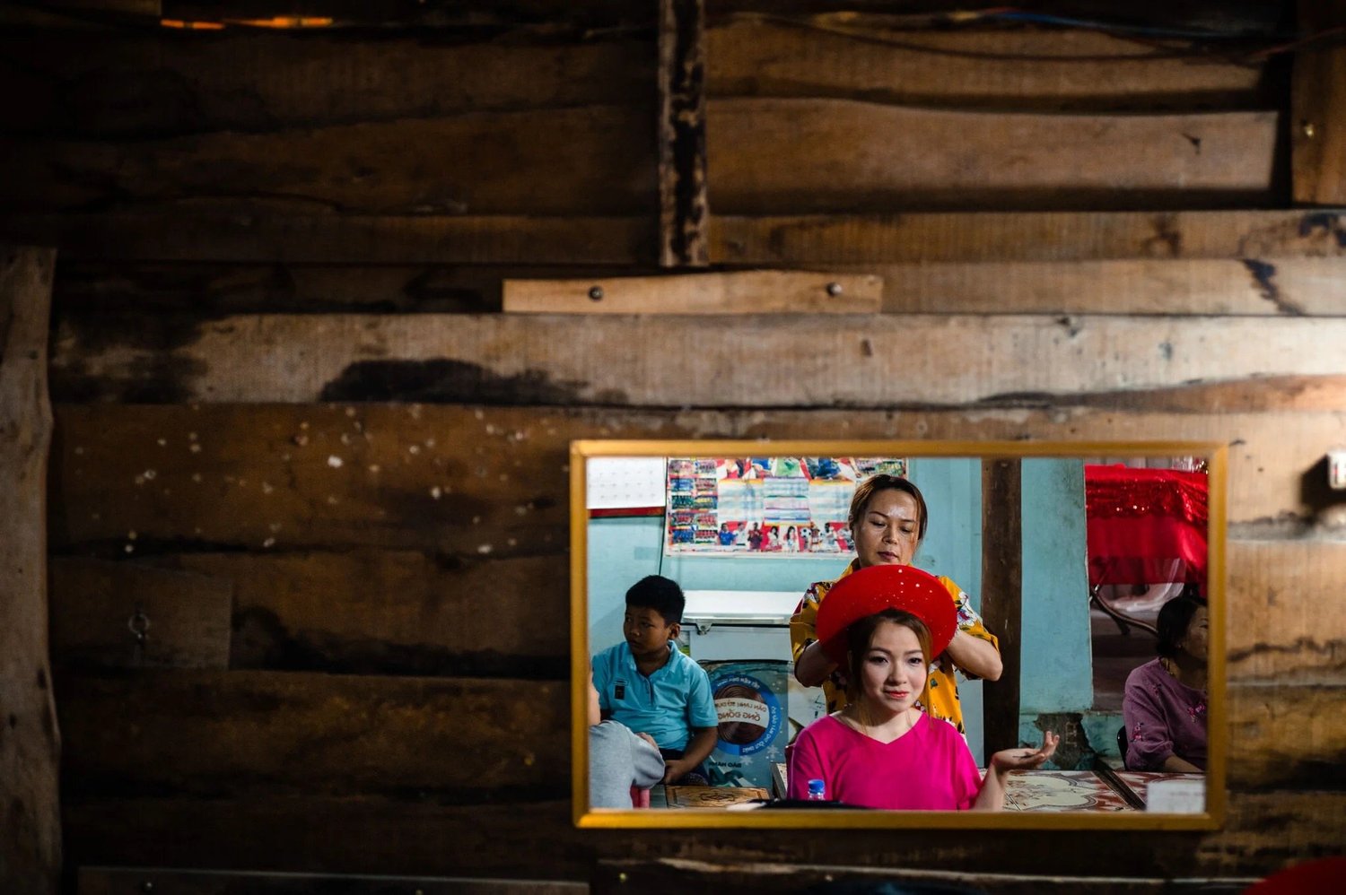 Hanh reflected in a mirror on a wooden wall while her headdress is adjusted