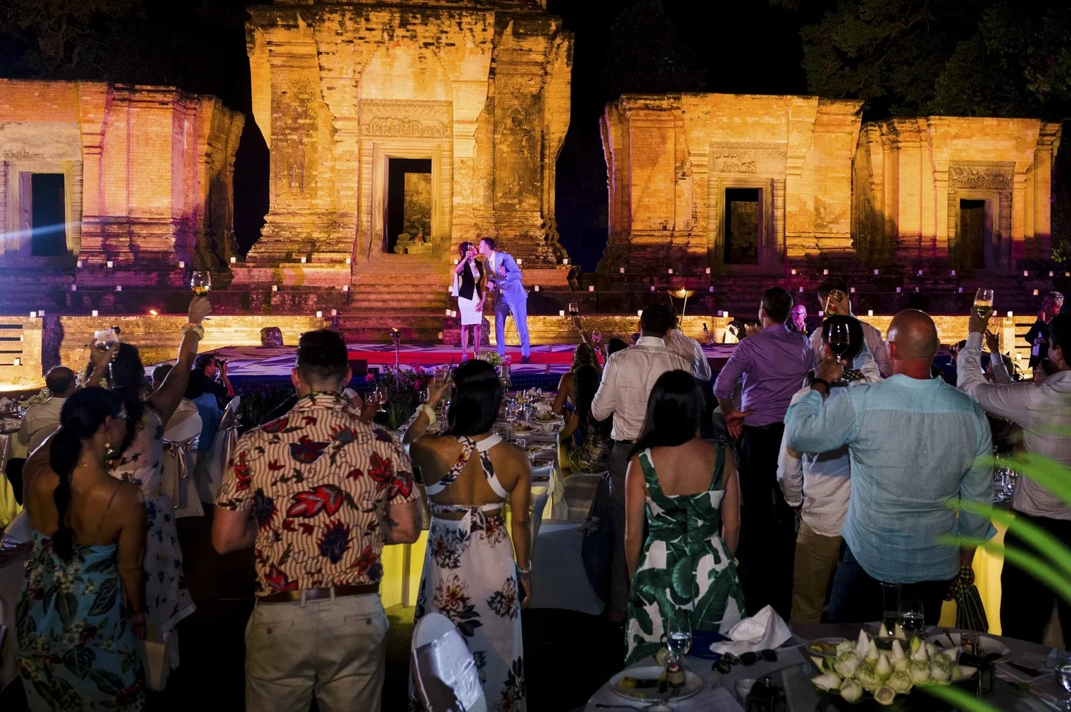Wedding guests raise their glasses towards Adam and Celeste in front of illuminated ancient temple towers in Cambodia.