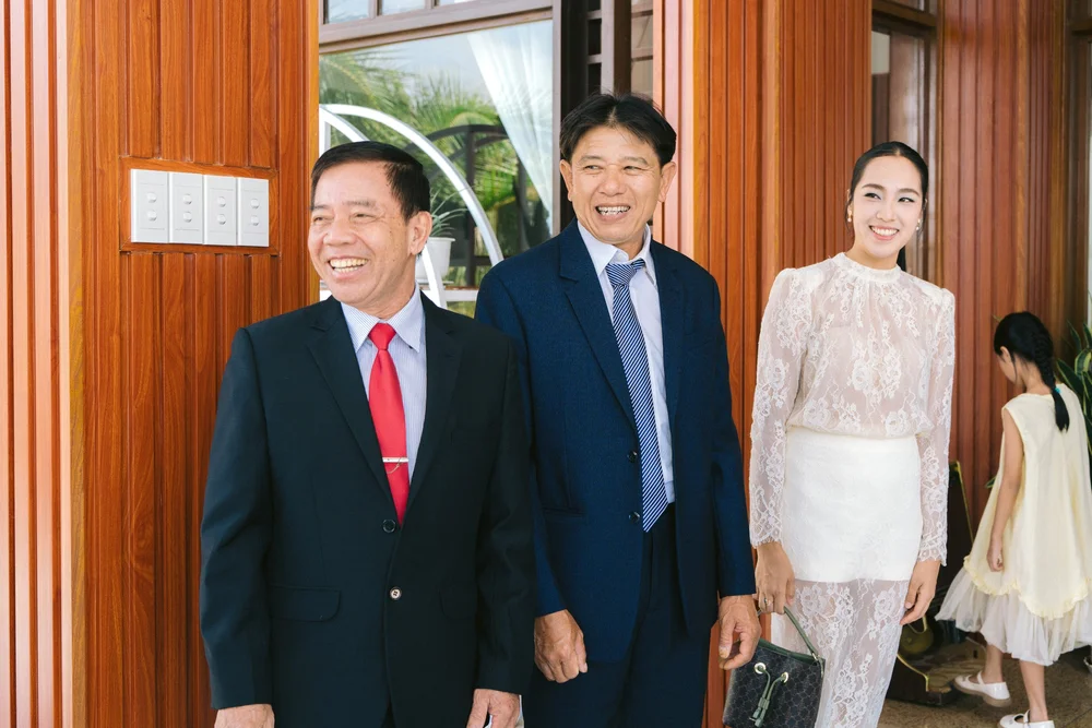 Two men in suits and a woman in a pale áo dài smiling at a doorway